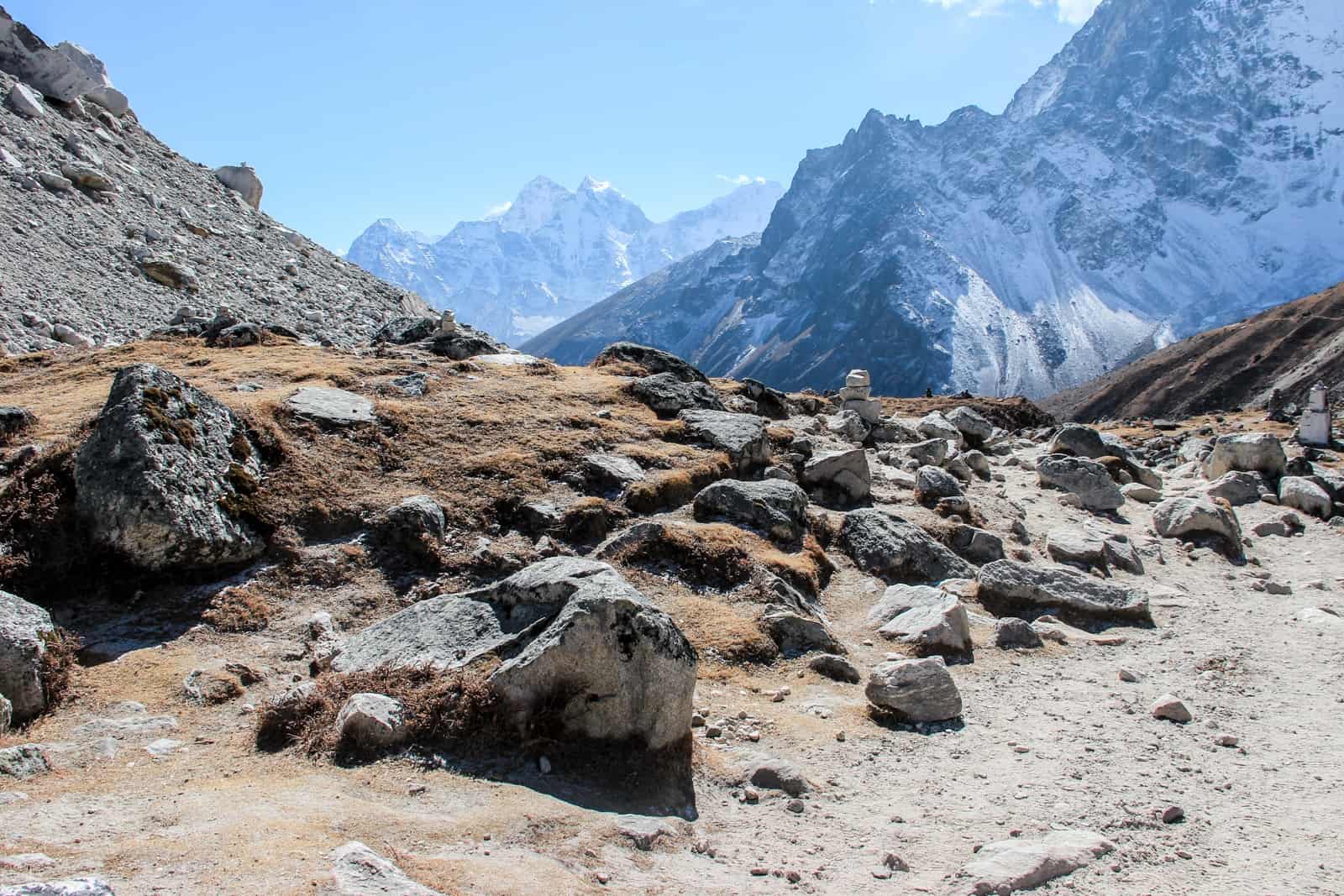 Large silver-grey rocks on golden brown, hilly soil, backed by silvery blue and white mountain peaks, on the Everest Base Camp trek route, 