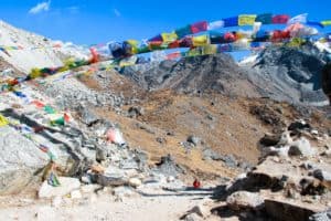 A trekker in a red jacket approaches a dusty path, surrounded by a panorama of sharp mountain peaks. The path is lined with giant rocks and a archway of multicoloured prayer flags hangs overhead.