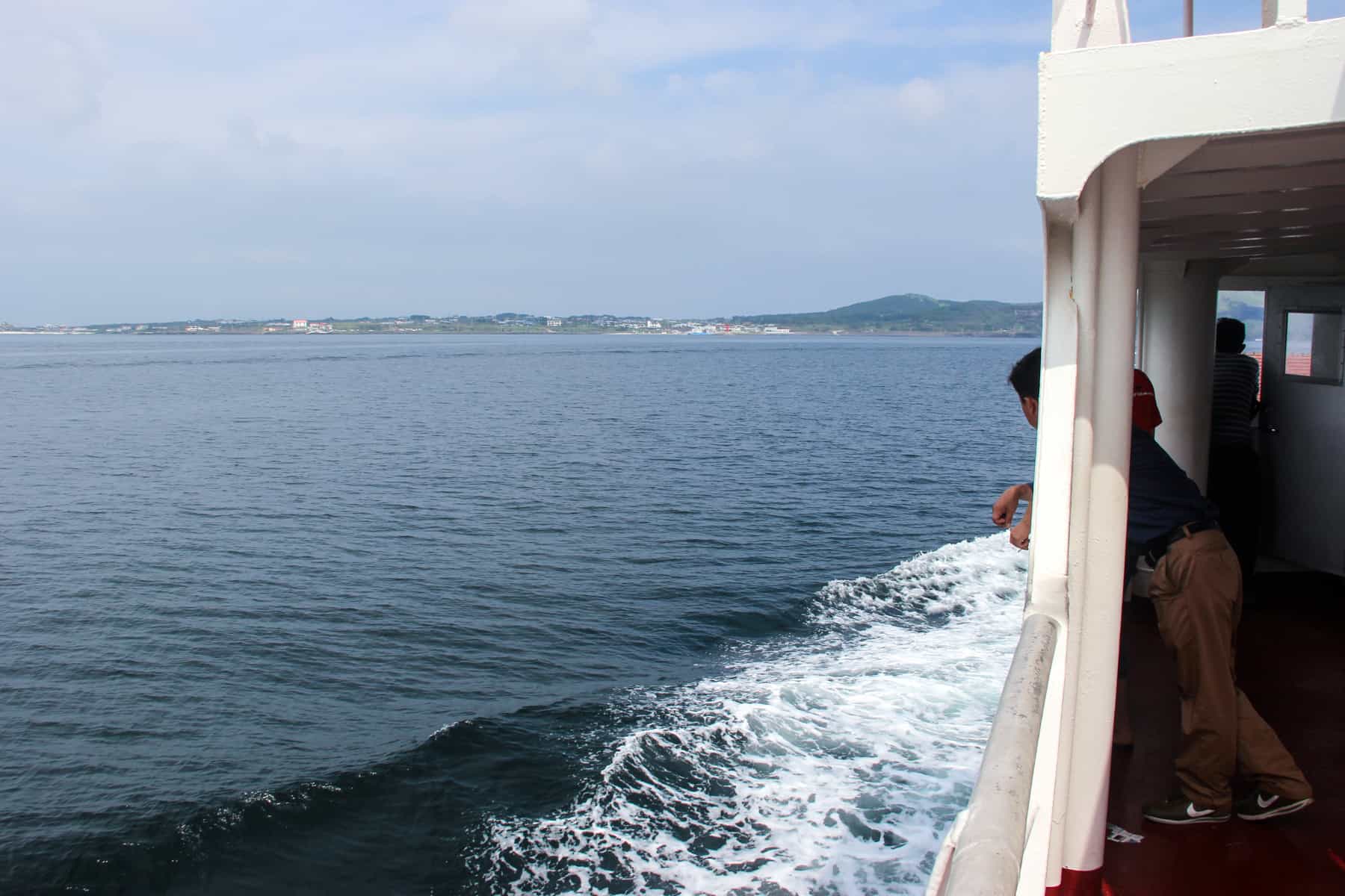 um homem inclina-se para fora do lado de um ferry branco para a Ilha de Jeju Coreia. O oceano é azul cinza e você pode ver os picos vulcânicos baixos da ilha no fundo.