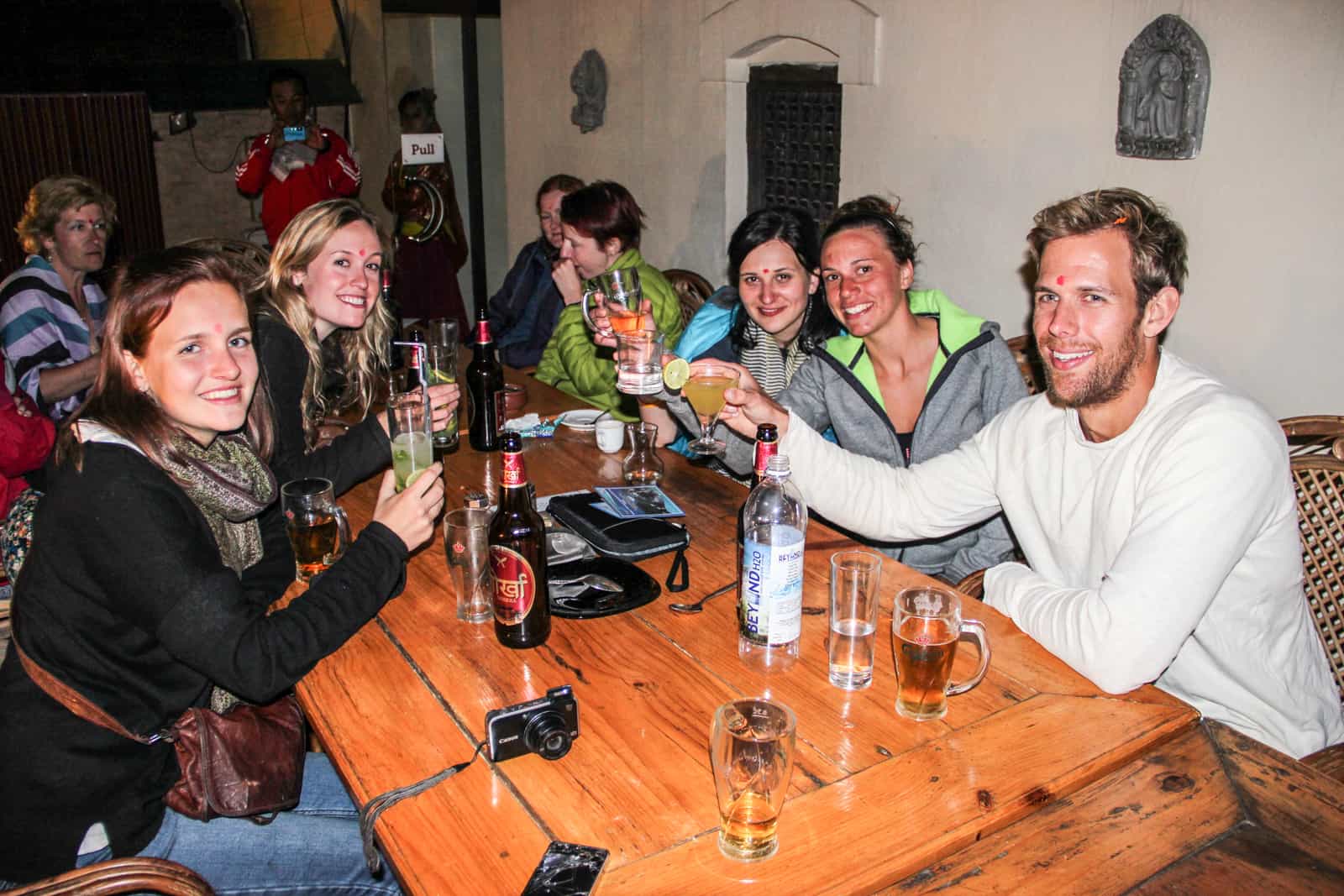 Four women and one man toast with glasses of drink at a golden brown wooden table 