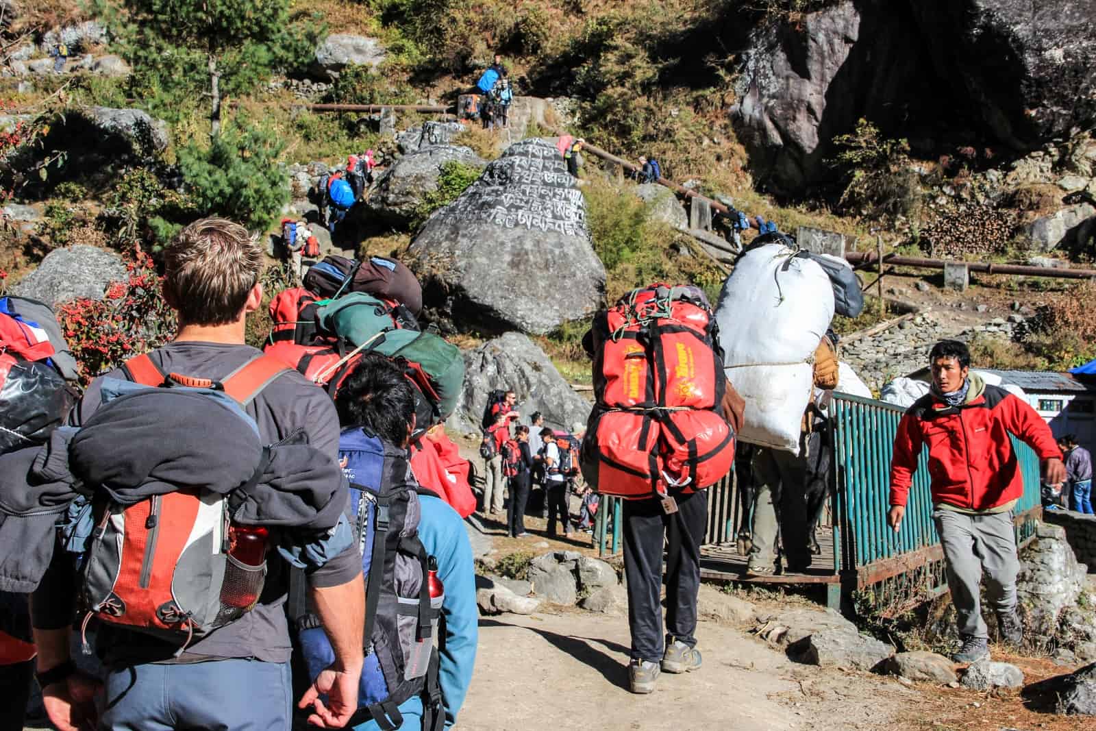 A line of trekkers and porters make their way across bridges and up the steep and rocky green hillside on the Everest Base Camp Trek