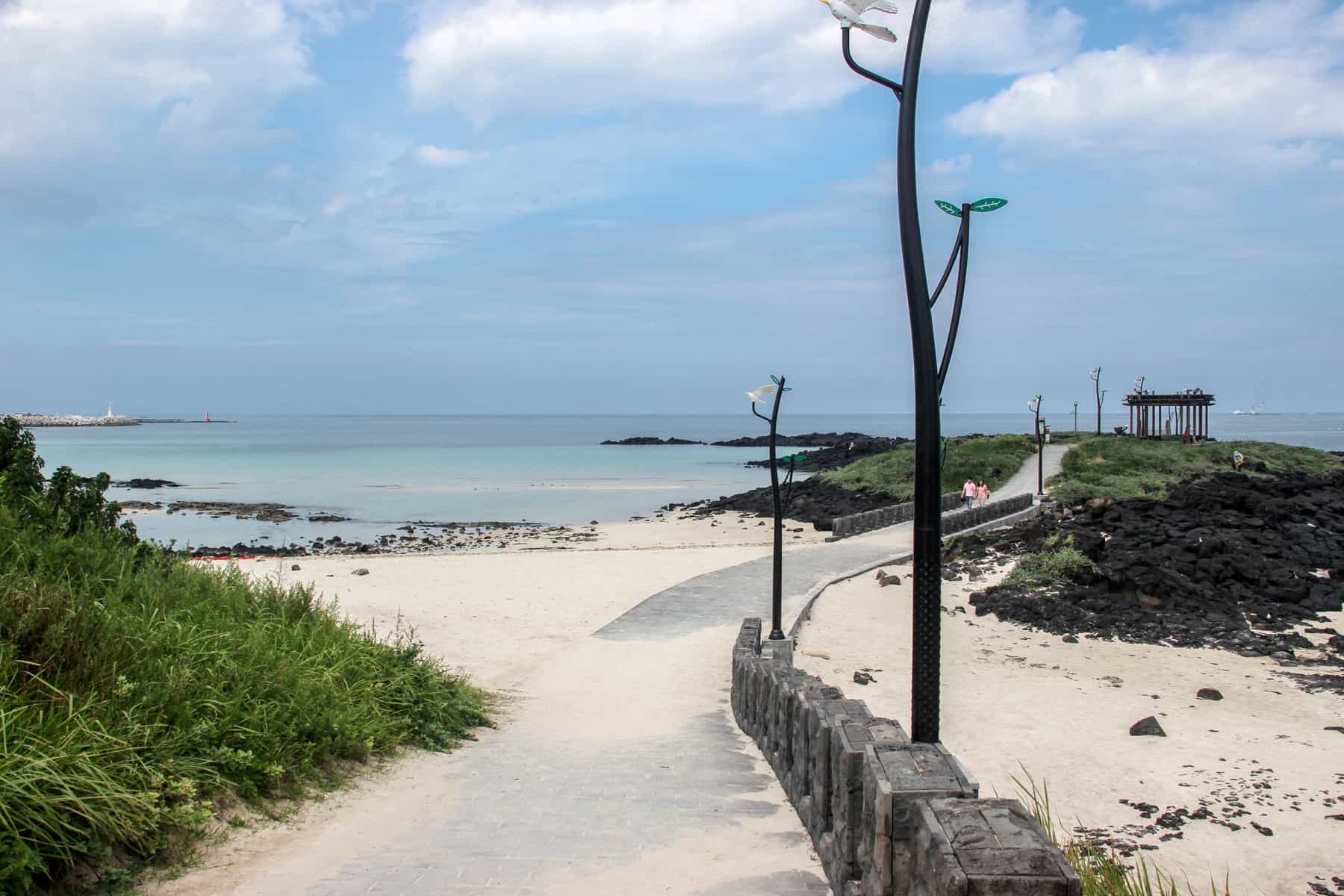 A sand covered pathway leads down to a white sand beach on Jeju Island. The water is a light turquoise blue and greenery is found next to the sand. 
