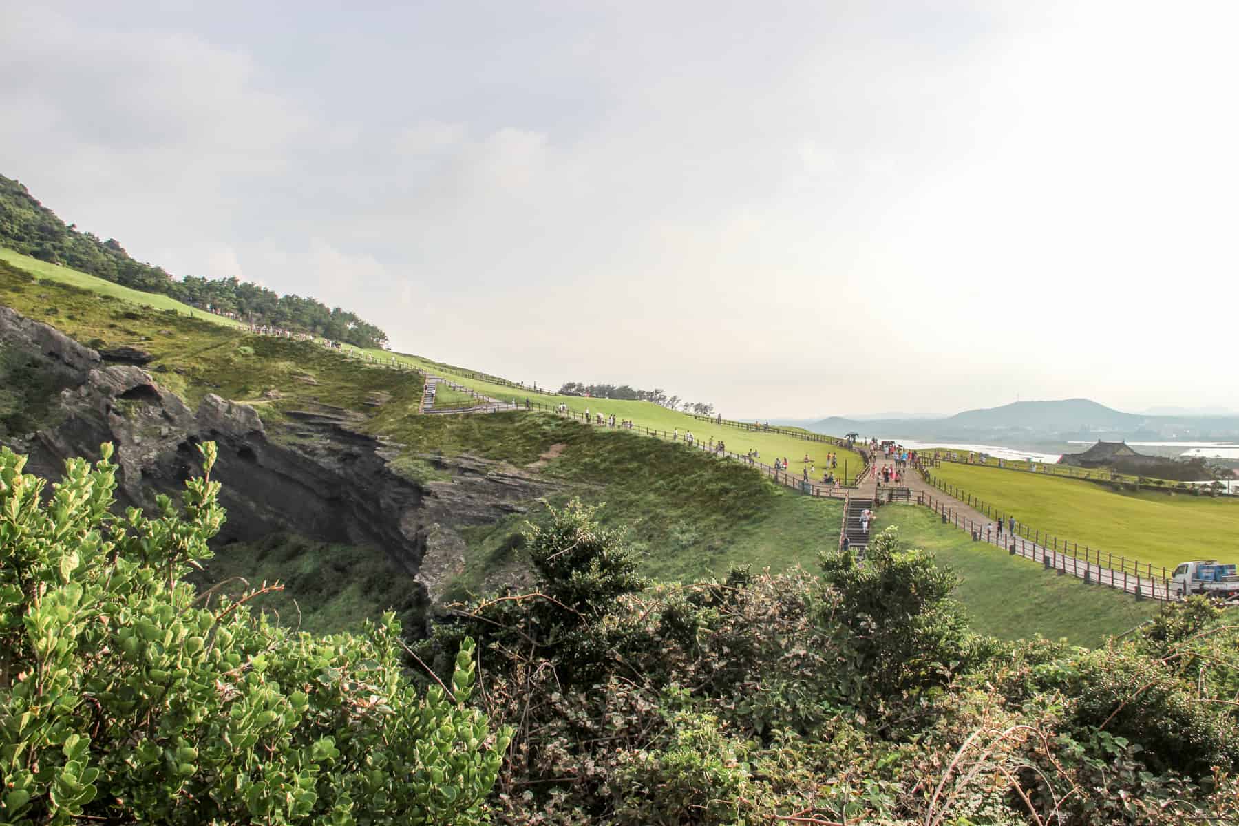 A view from the mountain pathway leading up Seongsan ilchulbong Peak on Jeju Island, looking down towards the coastline and a small mountain in the far ocean distance