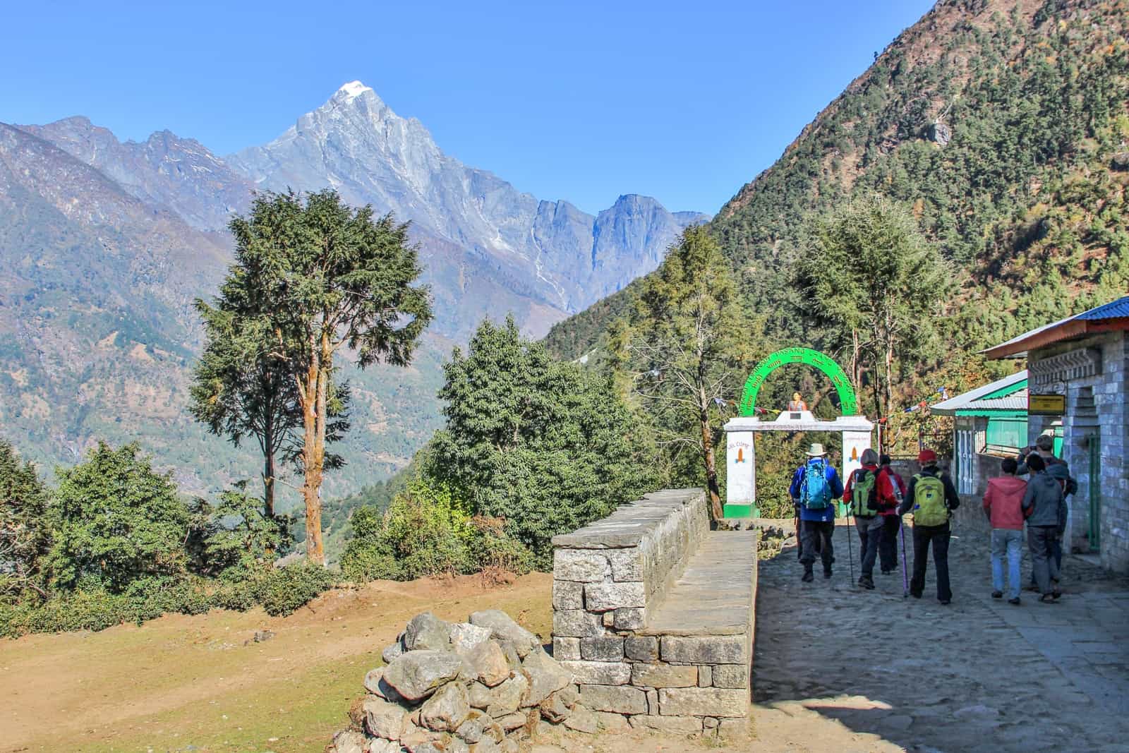 A small group of people walking on a stone path towards a white gate with a green arch - the starting gate of the Everest Base Camp trek in Lukla that looks towards the green forest and the Himalaya mountains