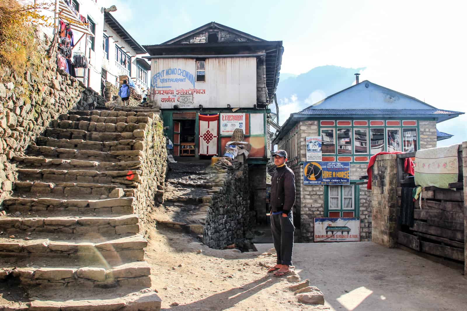 A man stands in front of two small houses with triangular rooftops and next to a sandy coloured staircase in the village of Namche Bazaar on the mountainous Everest Base Camp Trek route 