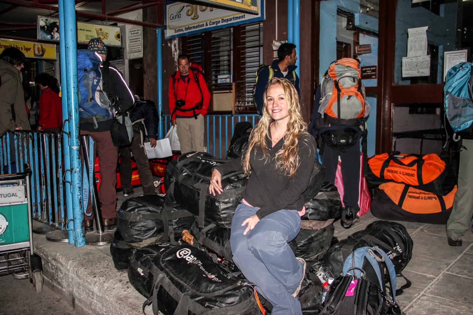 A woman sits on a pile of black trekking duffel bags outside a hotel entrance in Nepal. Other hikers can be seen in the background.