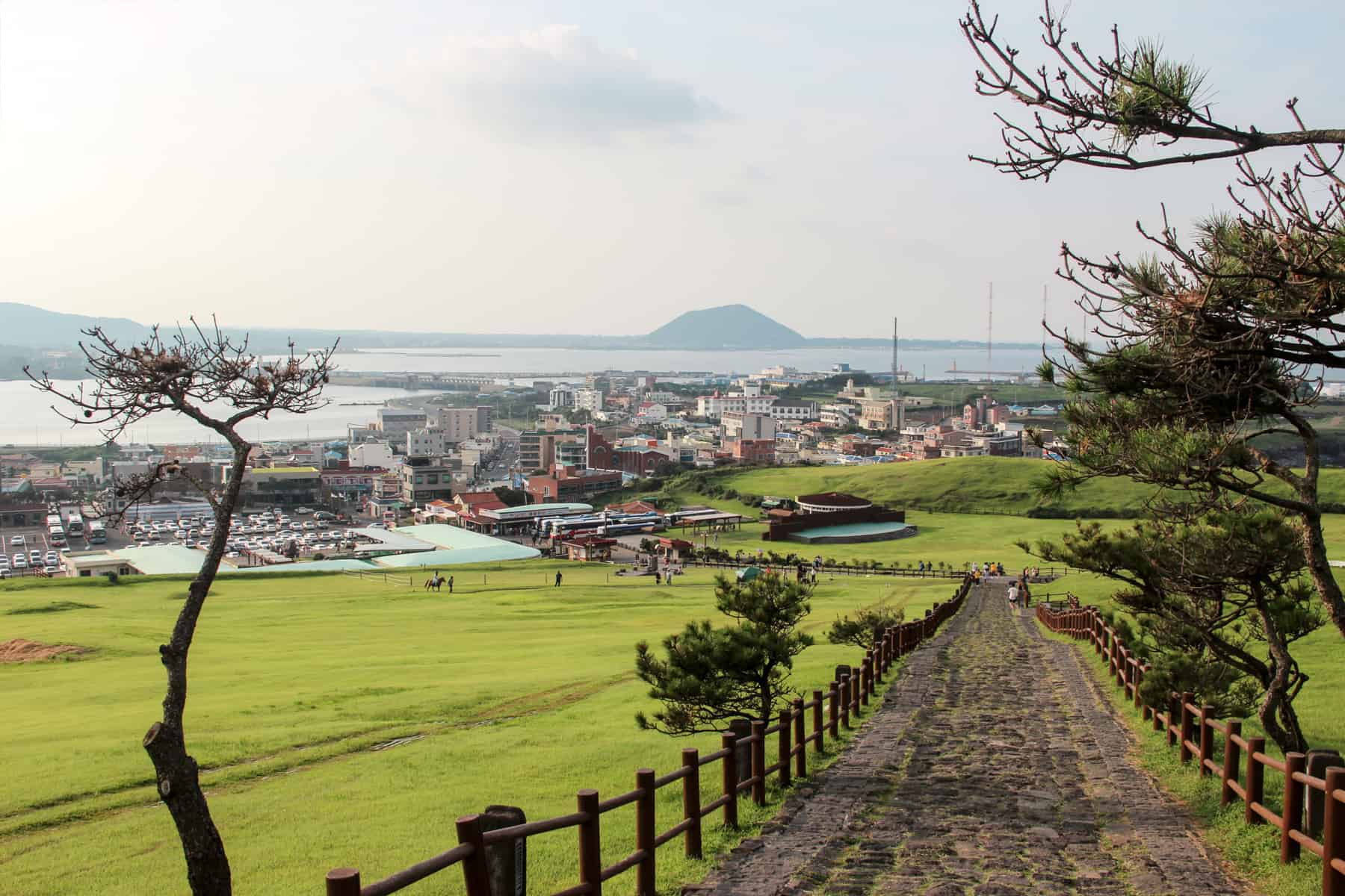 A view from the mountain pathway leading up Seongsan ilchulbong Peak on Jeju Island, looking down towards village buildinngs, the coastline and a small mountain in the far ocean distance