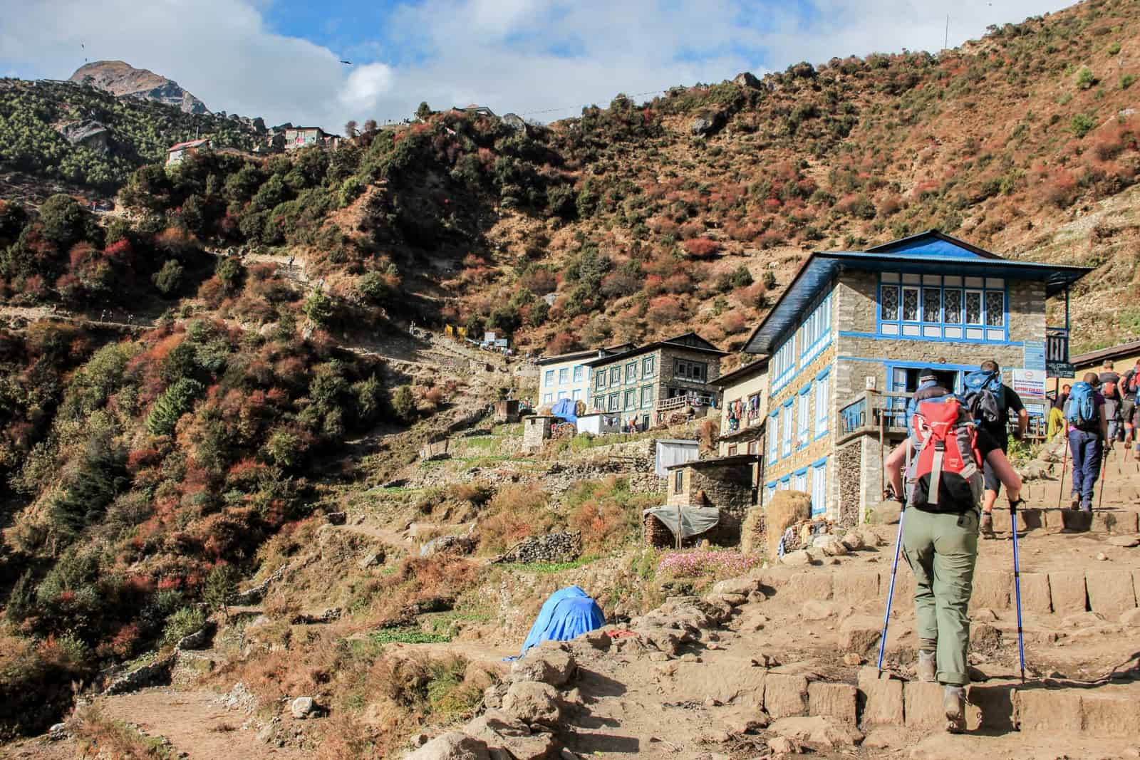 Three trekkers on an uphill climb on a steep worn trail past small huts on a orange rocky mountain face