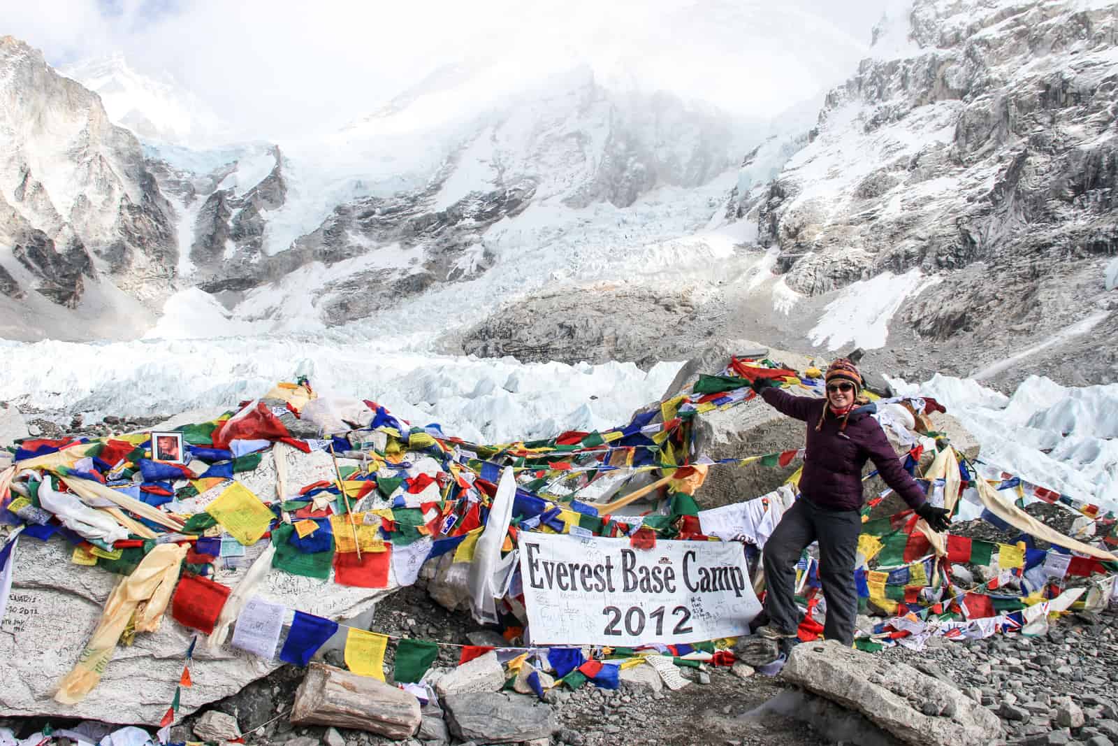 A woman in dark trekking clothing stands on Everest Base Camp in Nepal, on rocky ground surrounded by large chunks of ice and snow covered Himalaya mountains 
