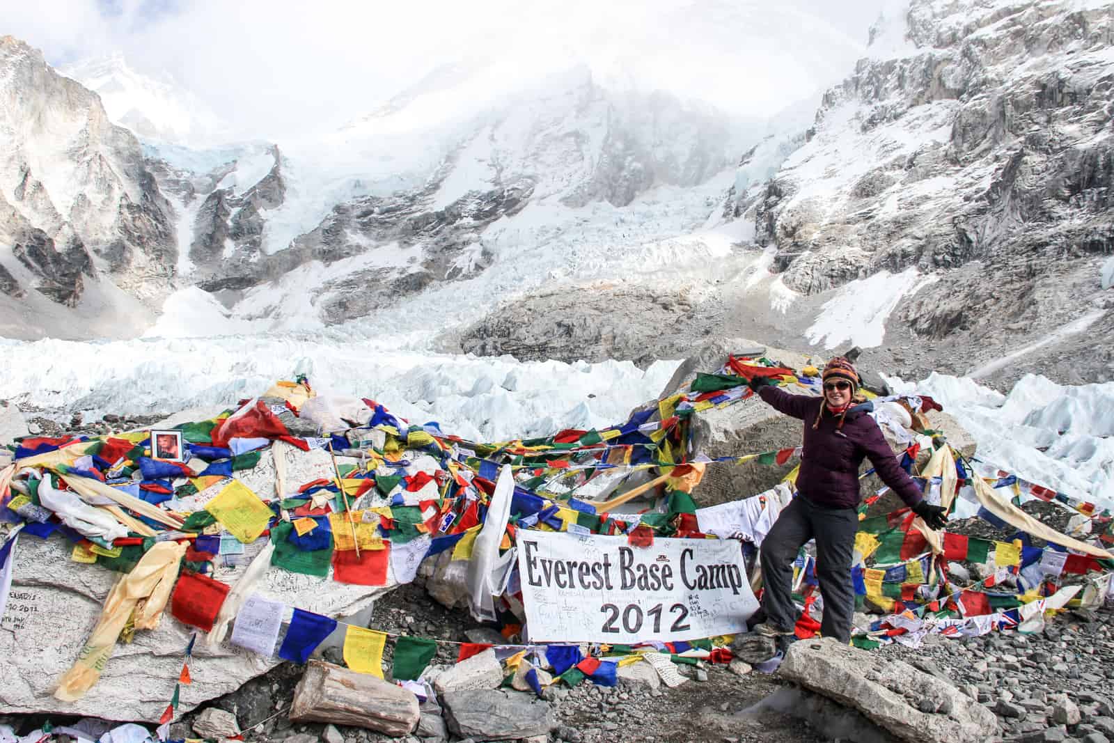 A woman in dark trekking clothing stands on Everest Base Camp in Nepal, on rocky ground surrounded by large chunks of ice and snow covered Himalaya mountains