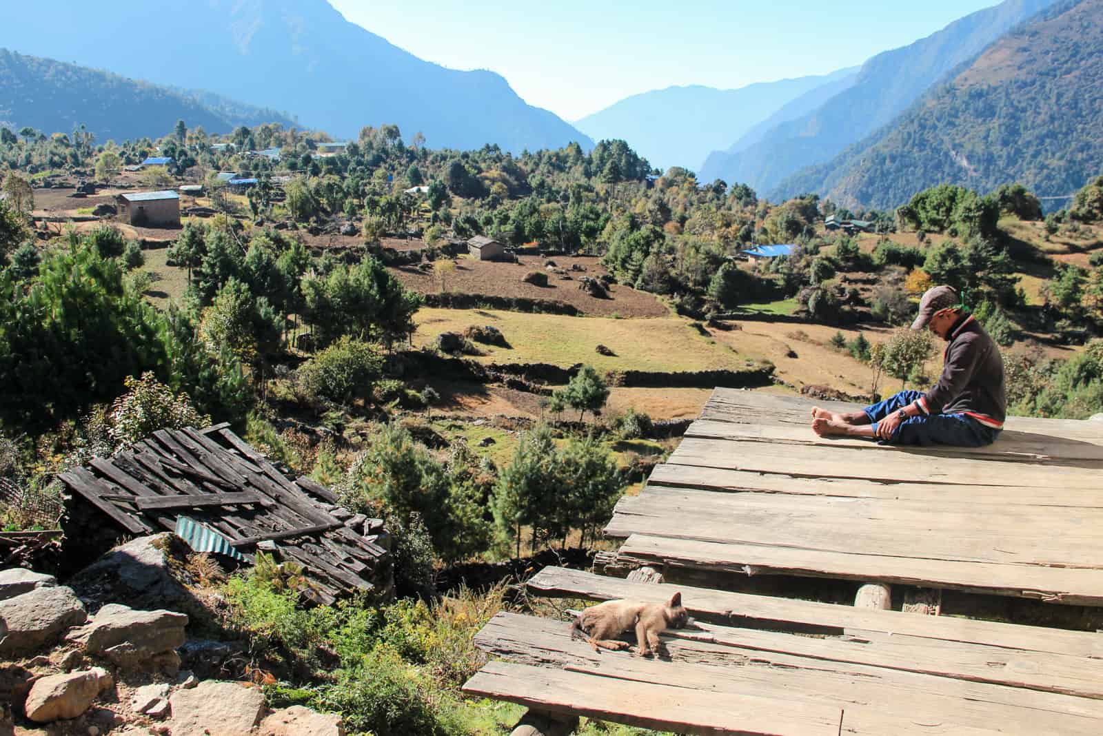 A man sits on a broken wooden platform with his dog, overlooking the green trees that fill Sagarmatha National Park on the Everest Base Camp Trek