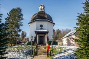 A woman in a red coat stands at the steps outside the small, white Silent Night Chapel in Oberndorf Village, Austria.