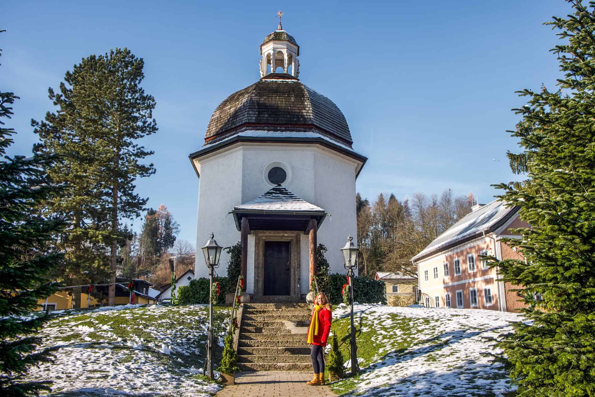 A woman in a red coat stands at the steps outside the small, white Silent Night Chapel in Oberndorf Village, Austria.