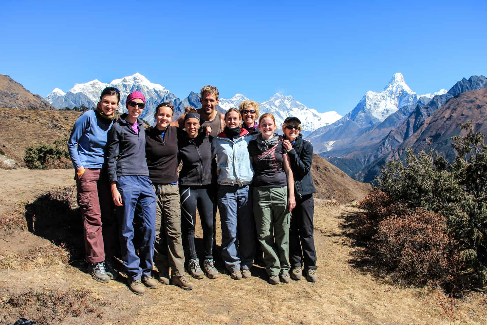 Nine people stand together on a dry, yellow mountain ridge in front of the snow capped Himalaya mountain range and Mount Everest