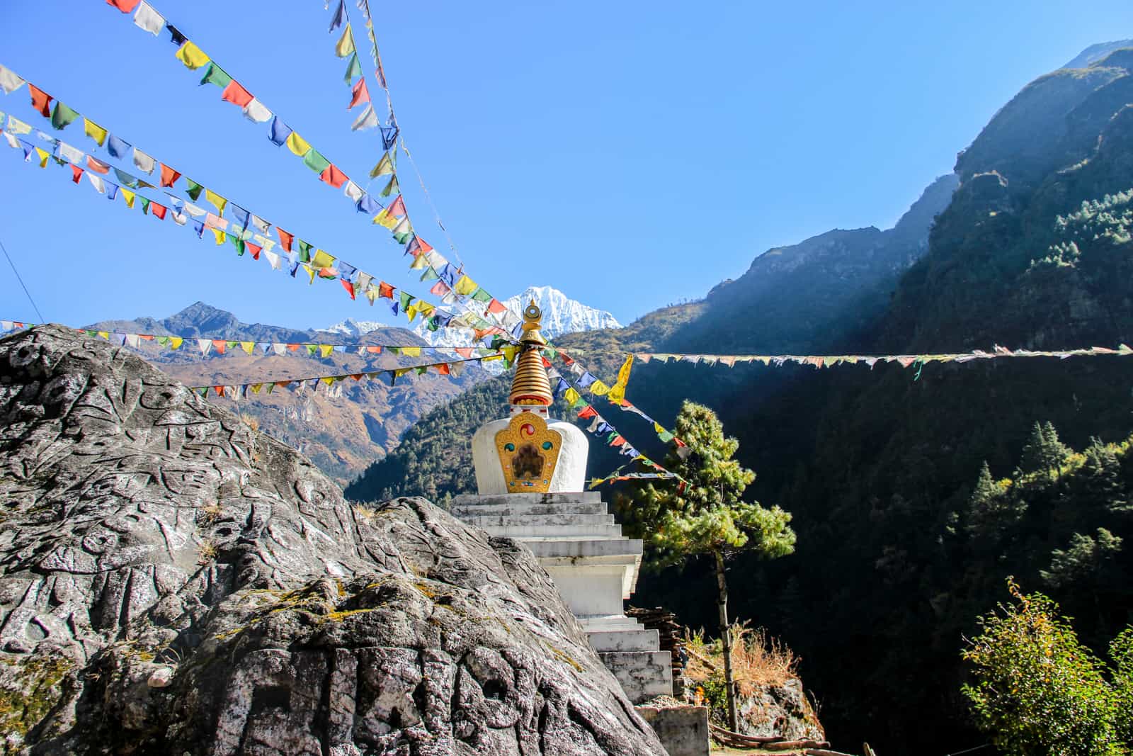 A small white temple structure with a golden spire sits on a rocks covered by lines of hanging multi-coloured prayer flags - a sacred site on the Everest Base Camp Trek