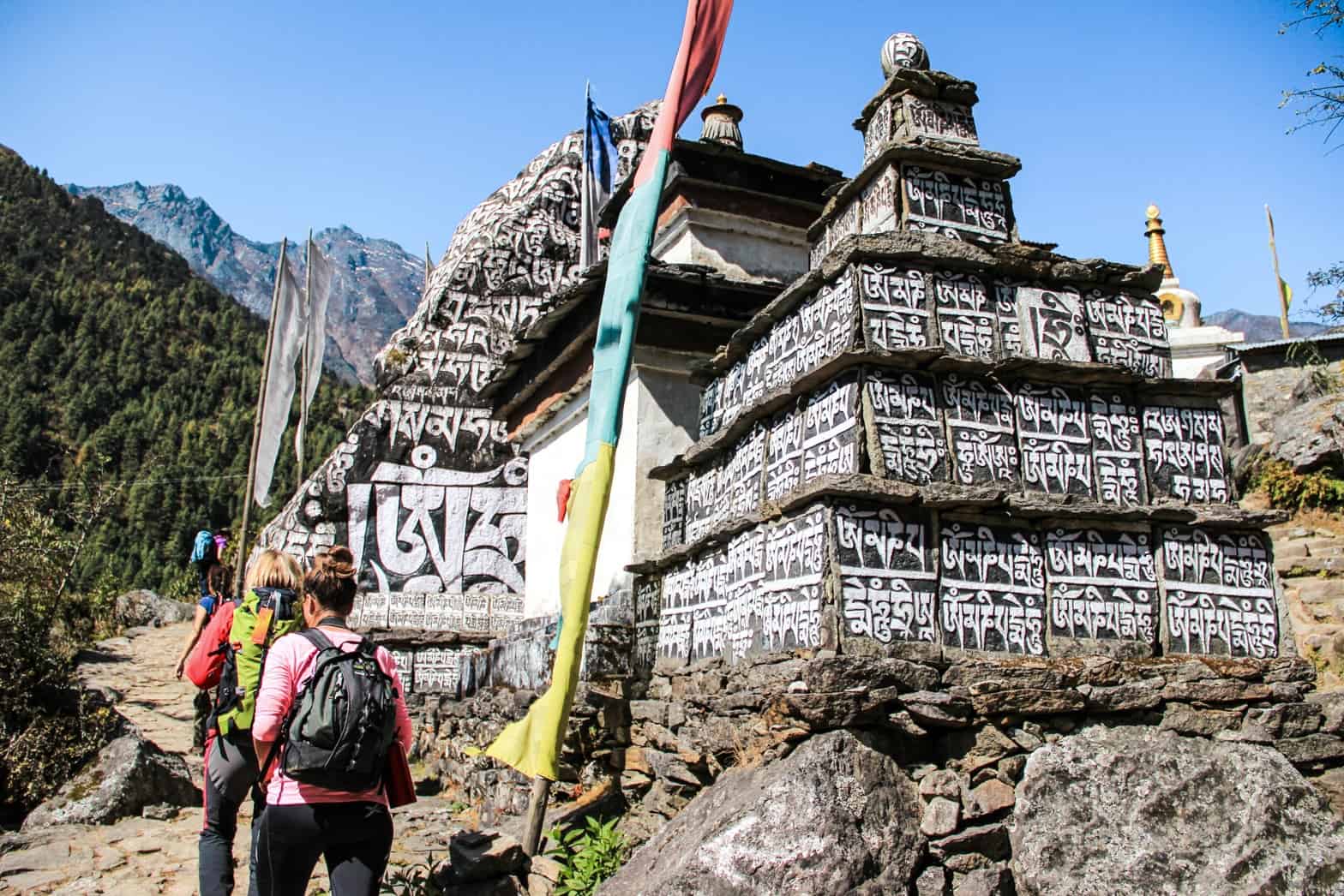 Hikers on the Everest Base Camp trek pass a sacred monument in the mountains - a tiered temple structure with black painted panels featured white painted script. Multi-coloured prayer flags hang next to the temple. 