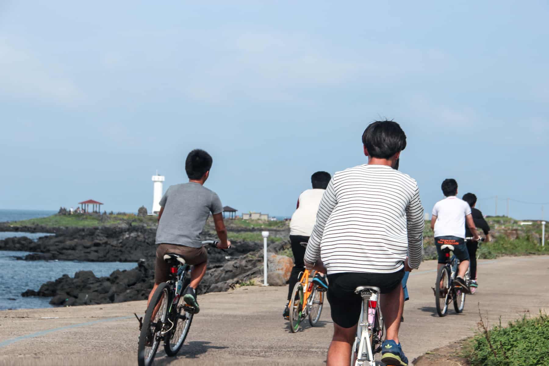 Five men in summer clothing ride bikes on a circular coastal pathway on Jeju Island, South Korea