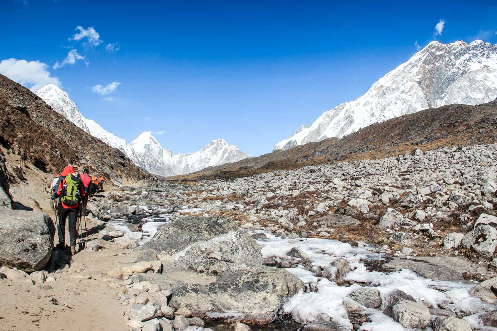 Two trekkers behind a line of yaks on a dusty path past a wide rocky scree on the Everest Base Camp Trek towards white mountain ranges. 