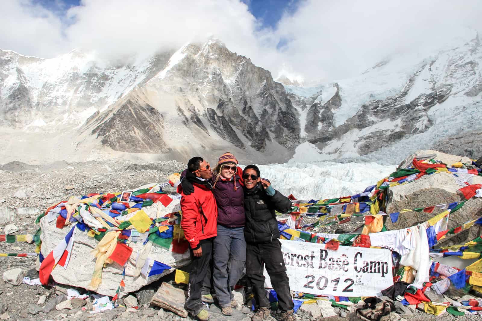 A woman in a purple jacket stands in the middle of two Nepali trekking guides at Everewst Base Camp. They stand in front of a rock mound in front of a text sign and prayer flags, and behind them is the white panorama of the mountain range.