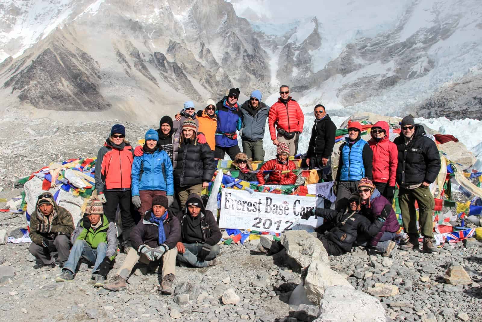 A group of trekkers at Everest Base Camp posing for a group shot around a sign with a 2012 date. Behind are the lower brown mountain slopes and a patch of ice.