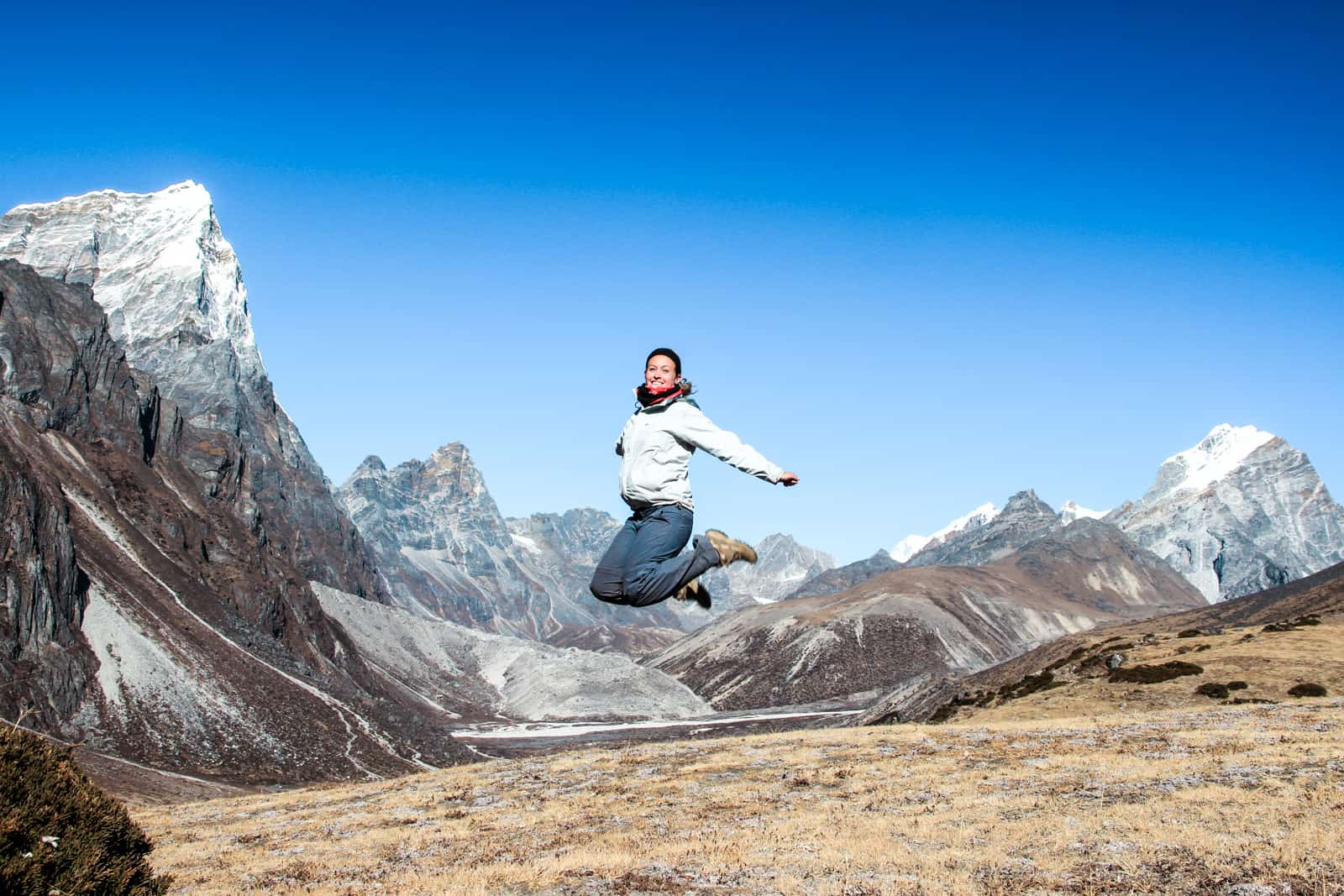 A woman in trekking gear jumps high to a background of rugged, pointed snow-capped mountain vistas on the Everest Base Camp trek