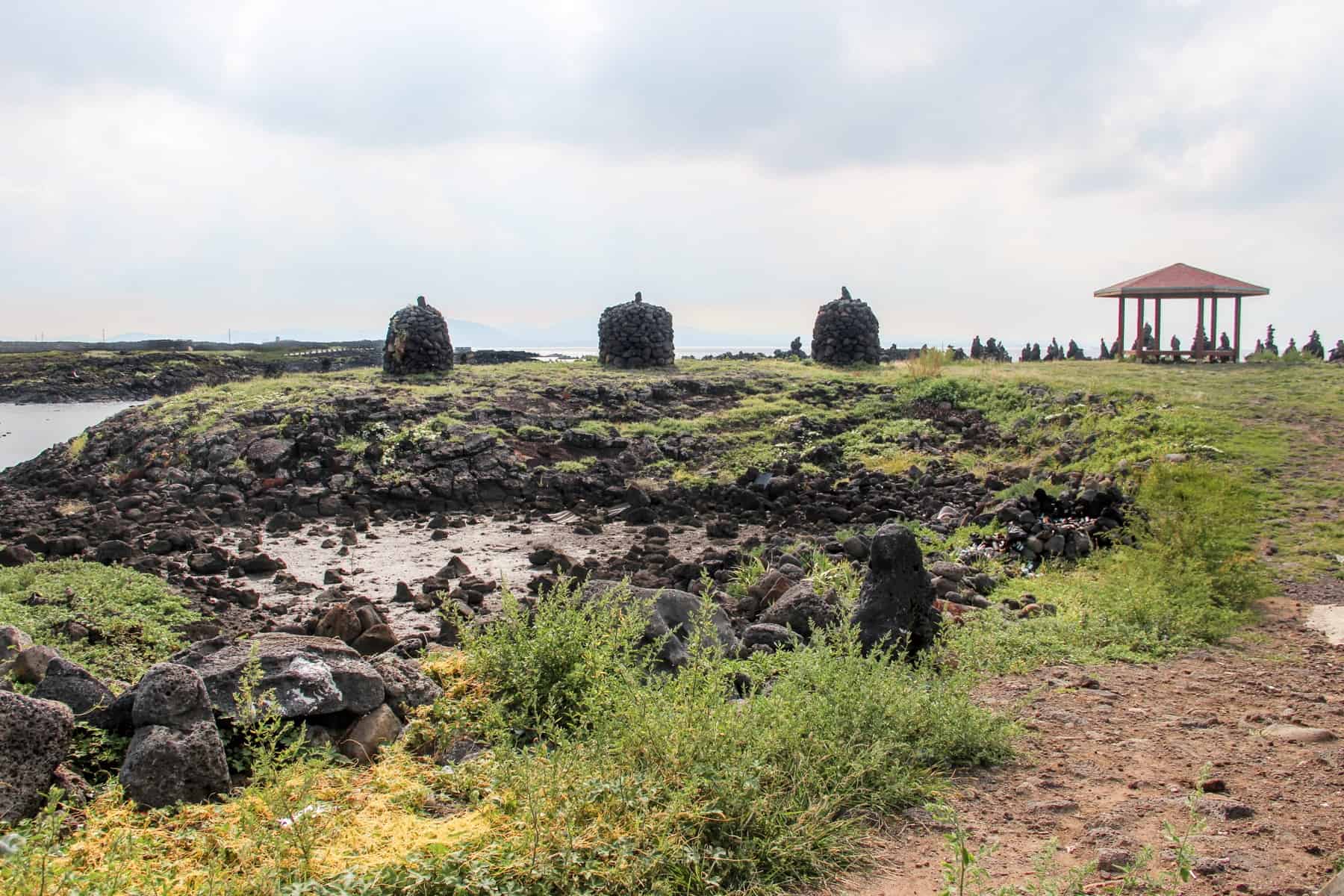 Three stone structures sit upon a rocky, green section of land that sits in the sea waters of Jeju Island in Korea