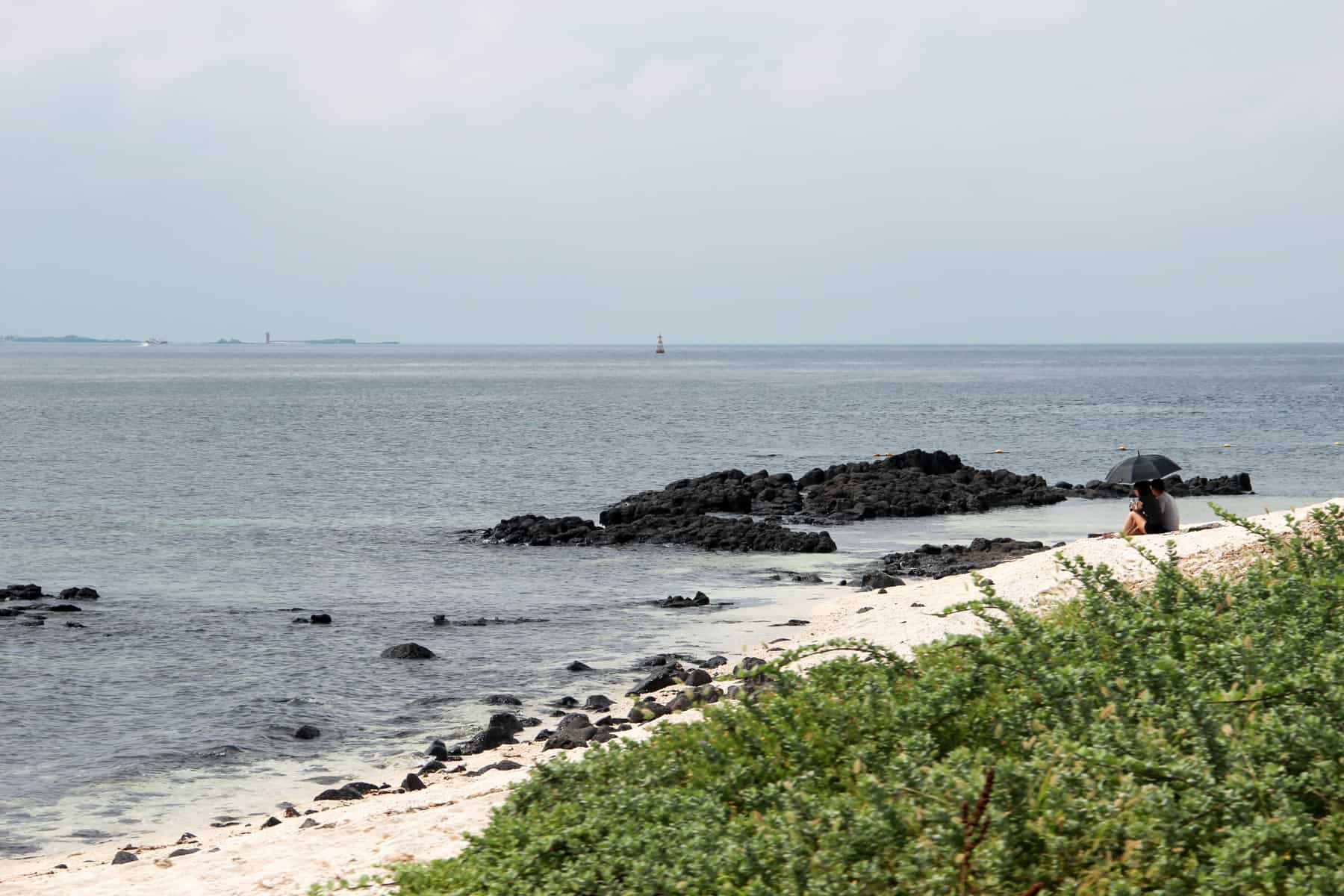 Uma praia de areia branca, próximo a um rock cheio de mar e apoiado por arbustos verdes, na Ilha Jeju, na Coreia do Sul