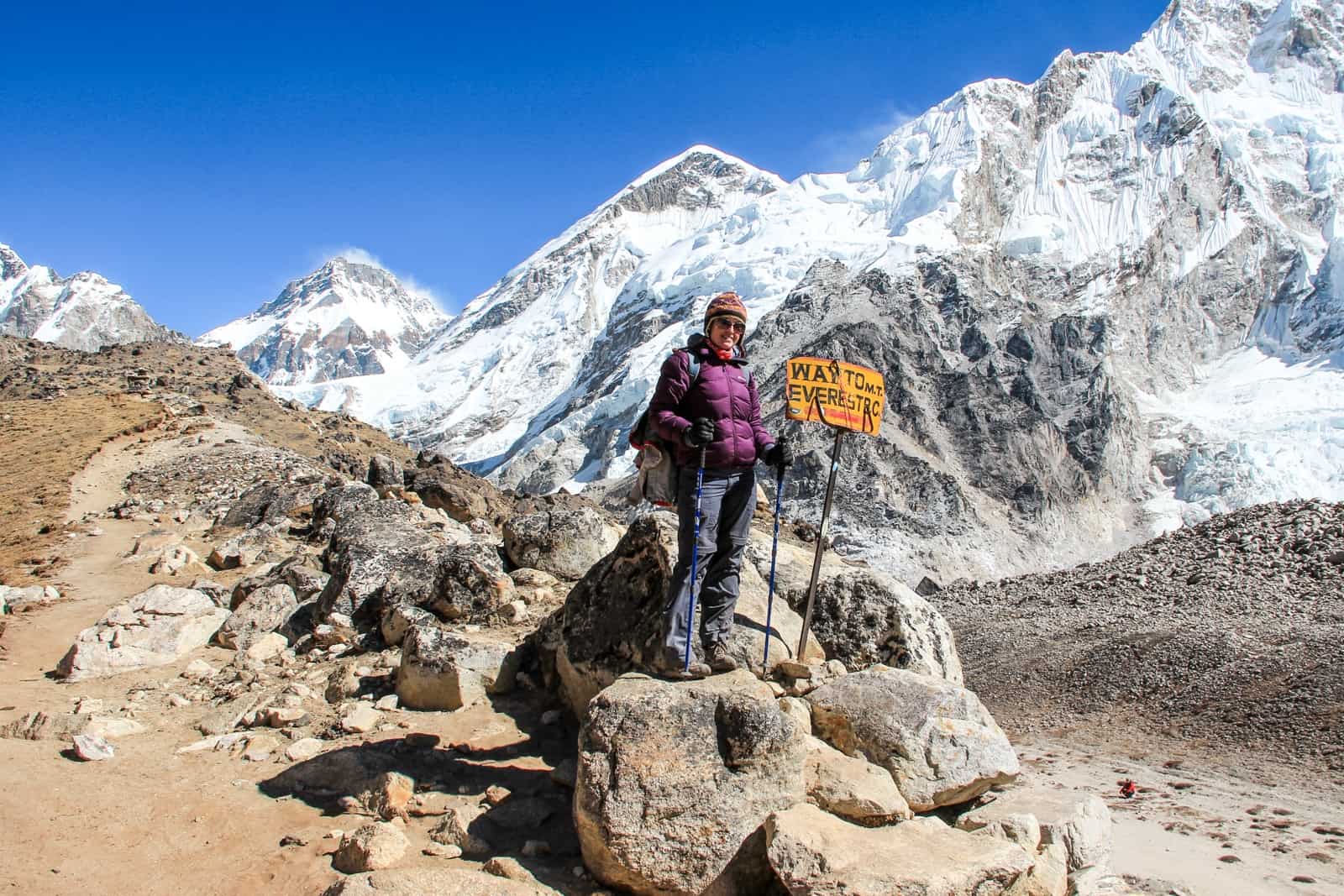 A woman in a purple jacket stands on a rock next to the famed "Way to Everest Base Camp" sign, backed by striking white mountains