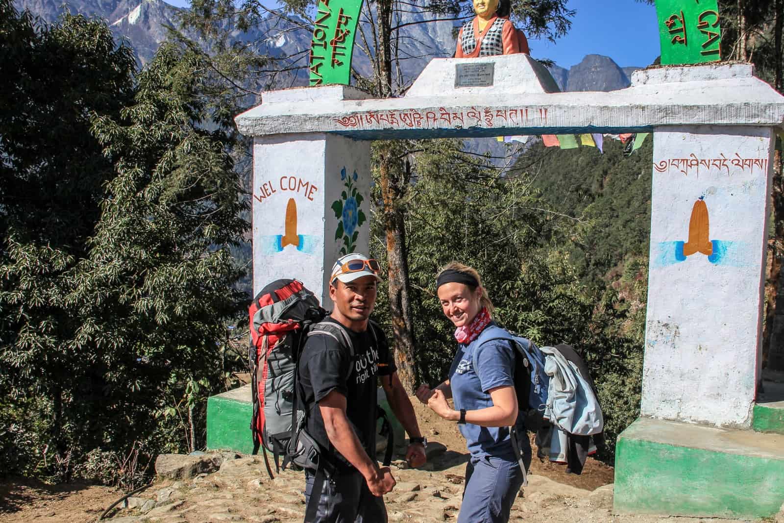 A Nepalese man and white woman stand outside of the 'Welcome' gate in Lukla on the EBC Trek and make arm muscle flex poses