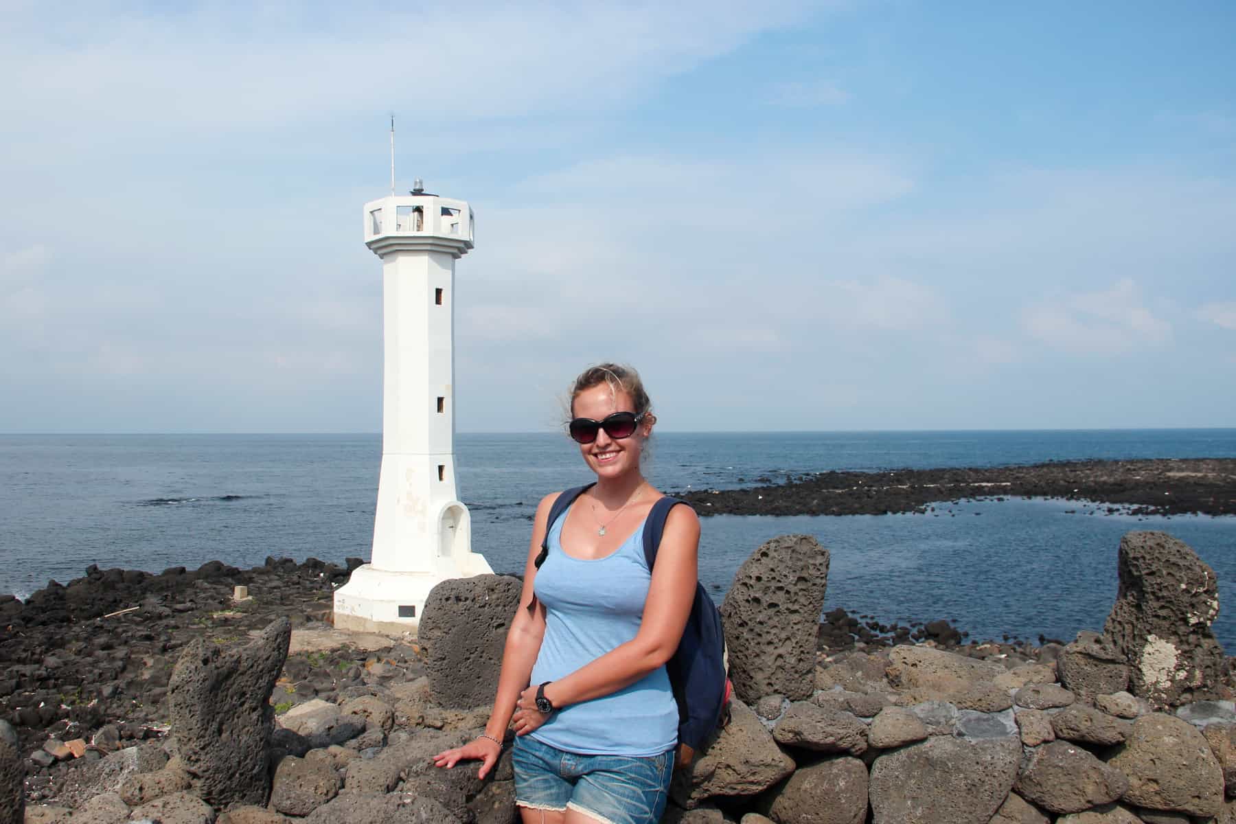 A woman in a blue vest and sunglasses stands in front of a White Lighthouse on the rocky Udo Maritime Park on Jeju Island in Korea