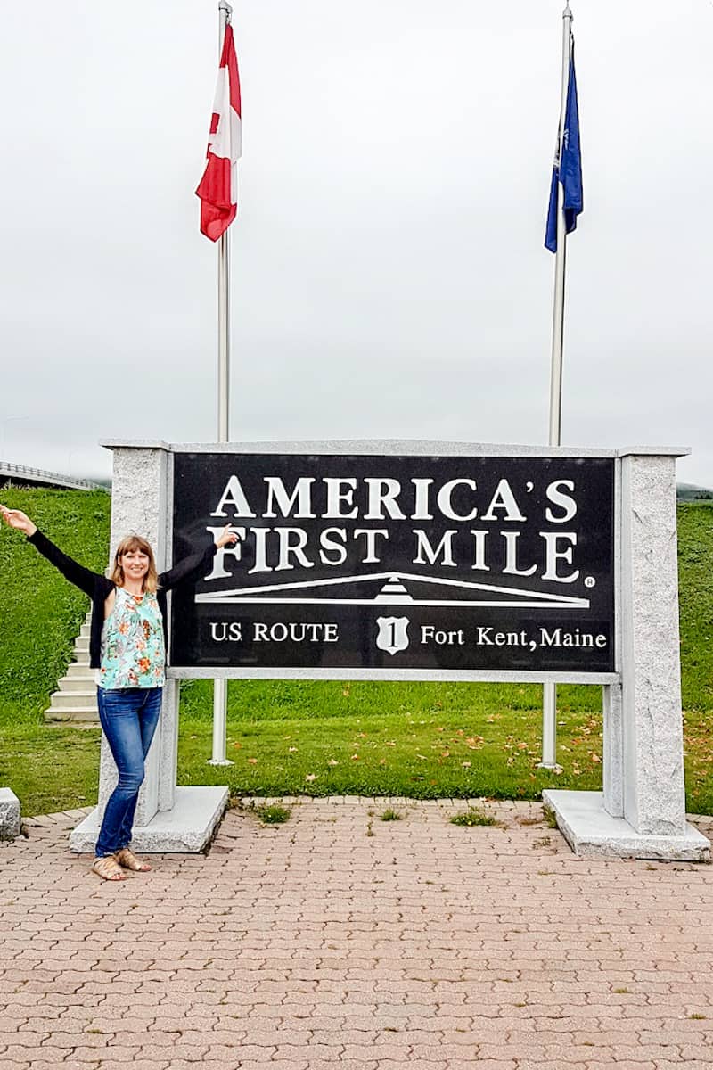 A woman stands in front of a sign topped with two flags, which reads: America's First Mile US Route 1 Fort Kent, Maine.