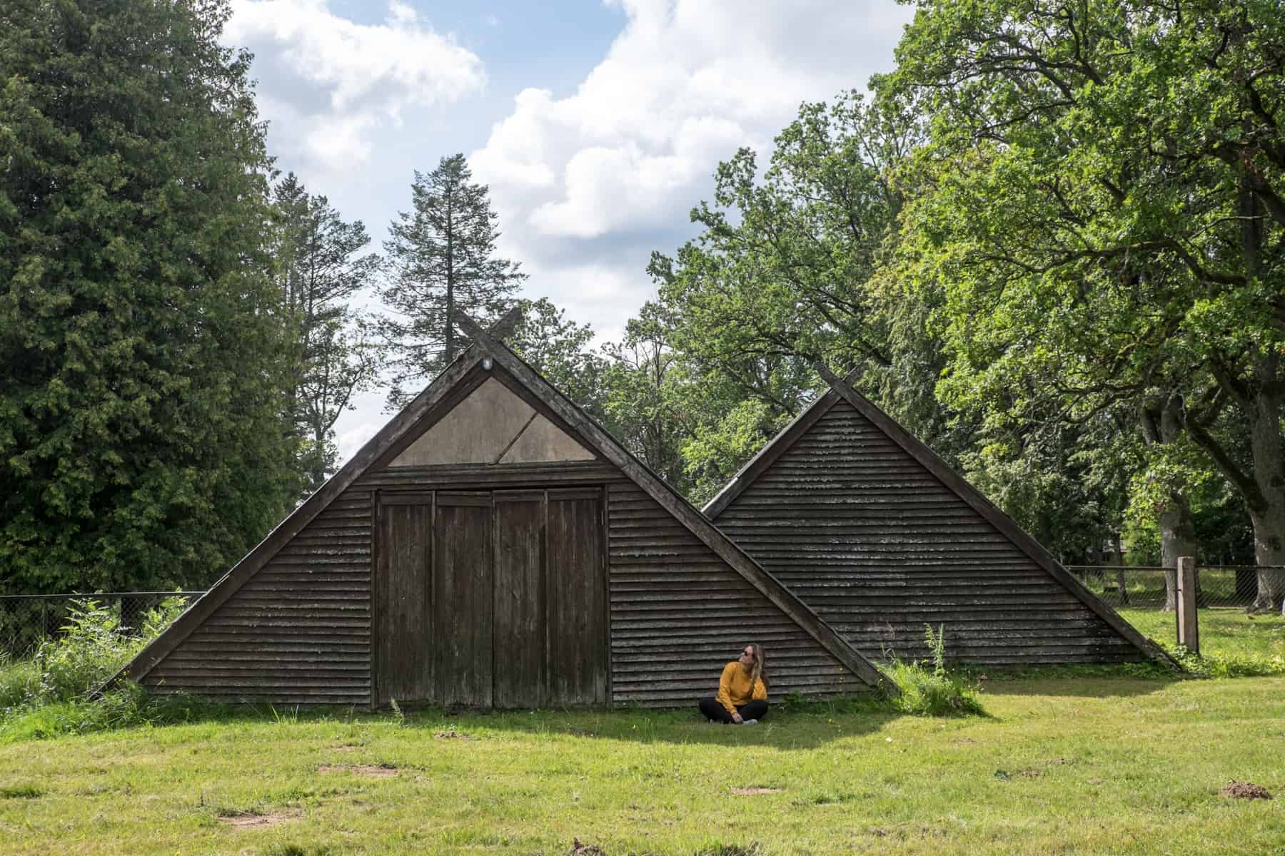 A woman in a yellow jumps sits on the grass in front of a brown, wooden triangular building, in the parkland of Sigulda, Latvia. 