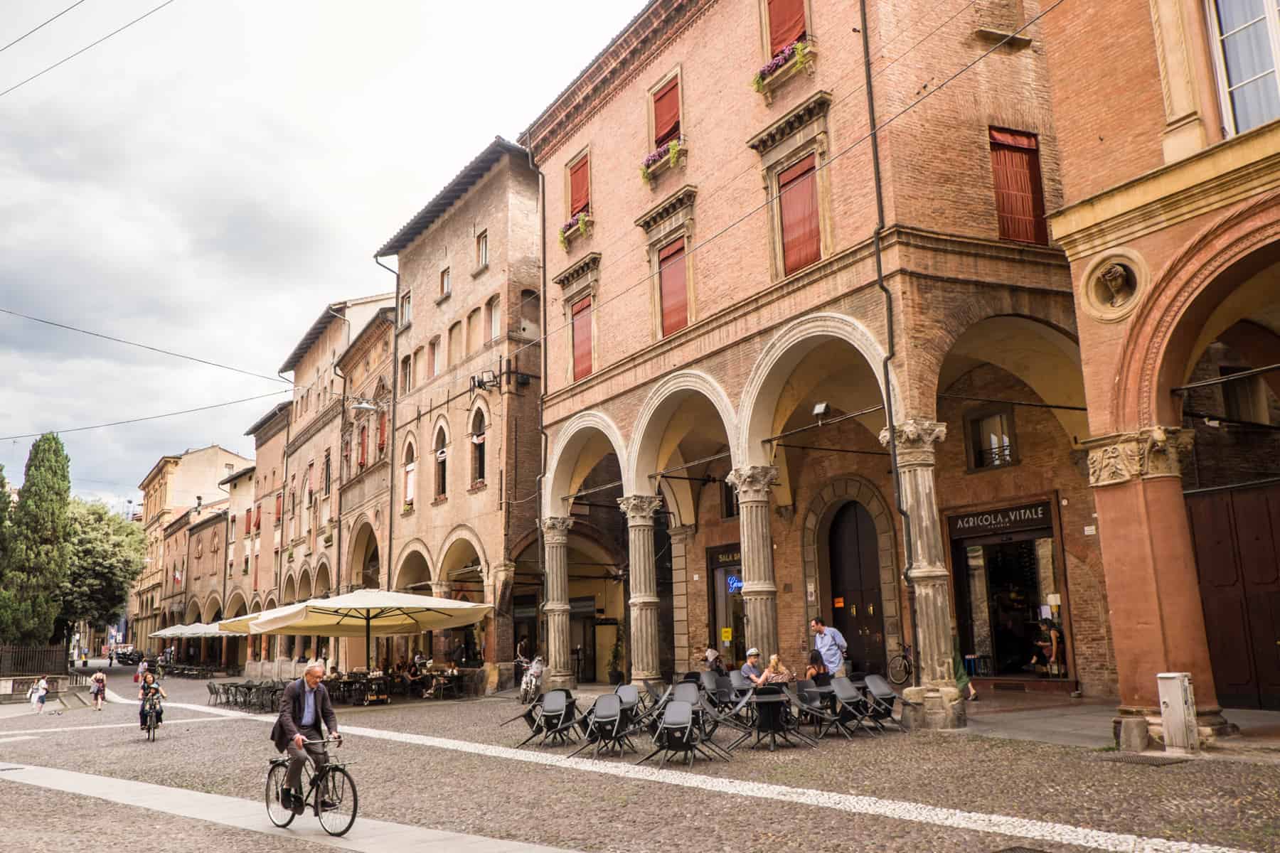 People sit at a cafe under umbrellas and a man rides a bike past opulent white arched orange buildings in Bologna, Italia. 
