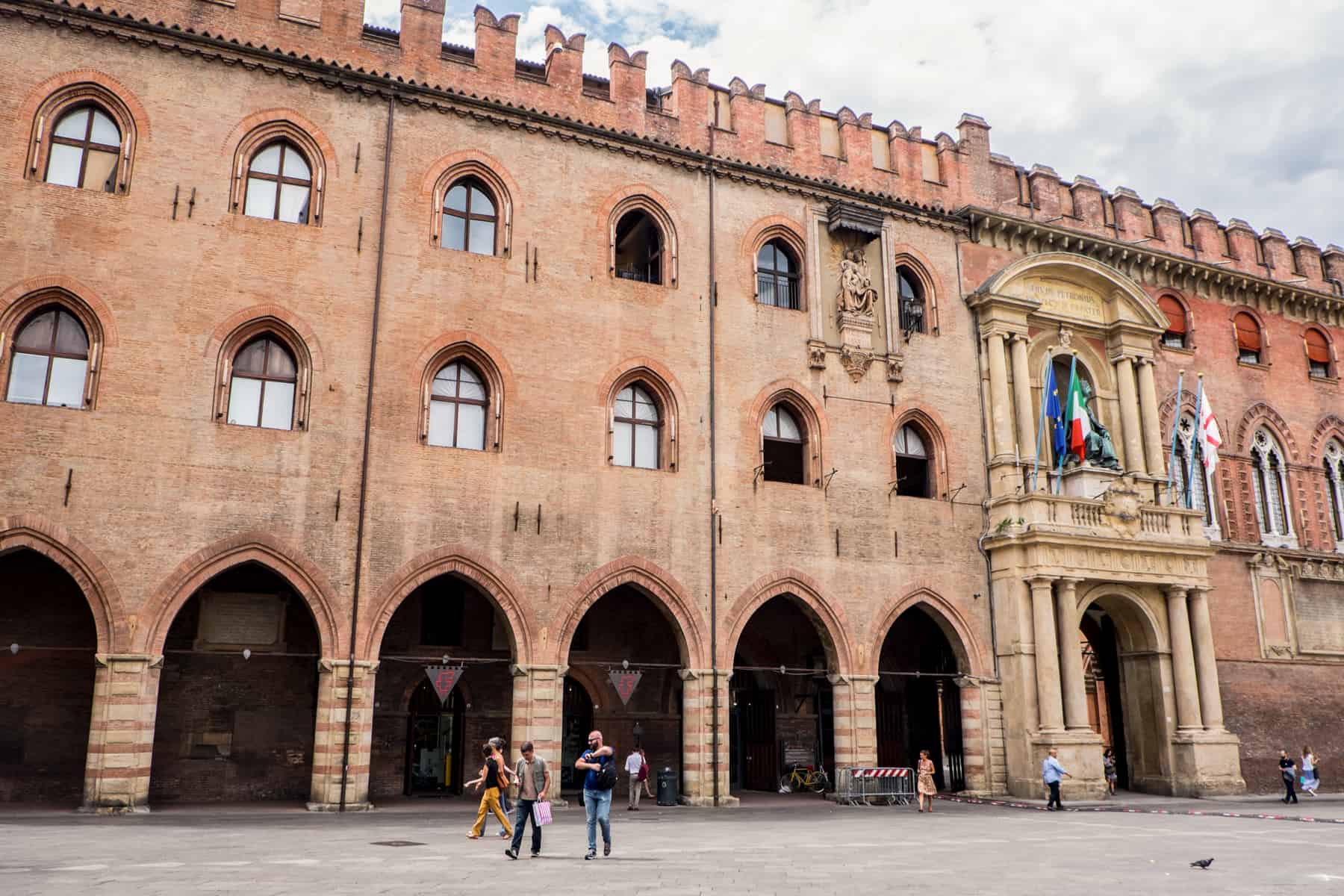 A long orange-brown castle looking structure with archways, rooftop turrets and a yellow-stone columned entrance door is the oldest university in the world in Bologna.