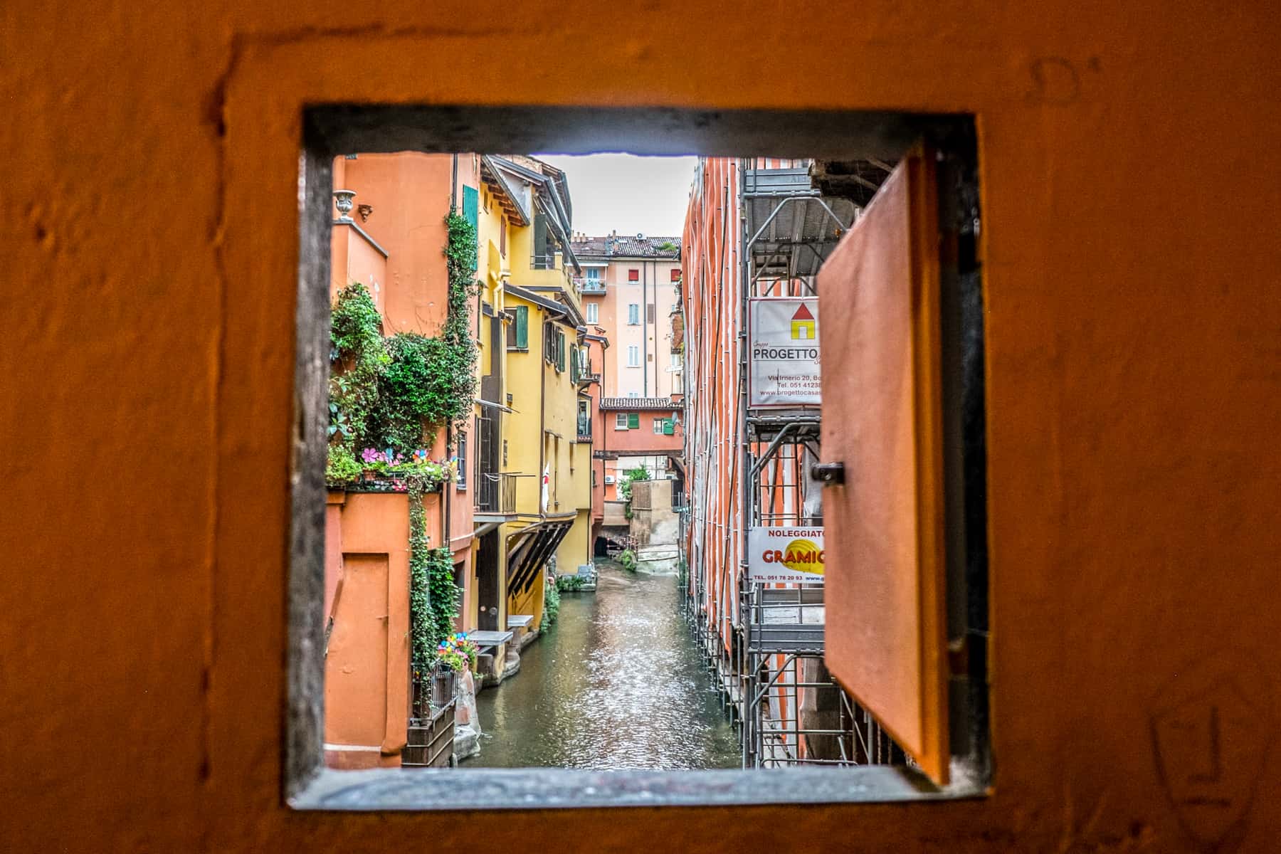 A window on a ochre orange wall with a view to a small canal in Bologna, lined with orange and yellow buildings. 