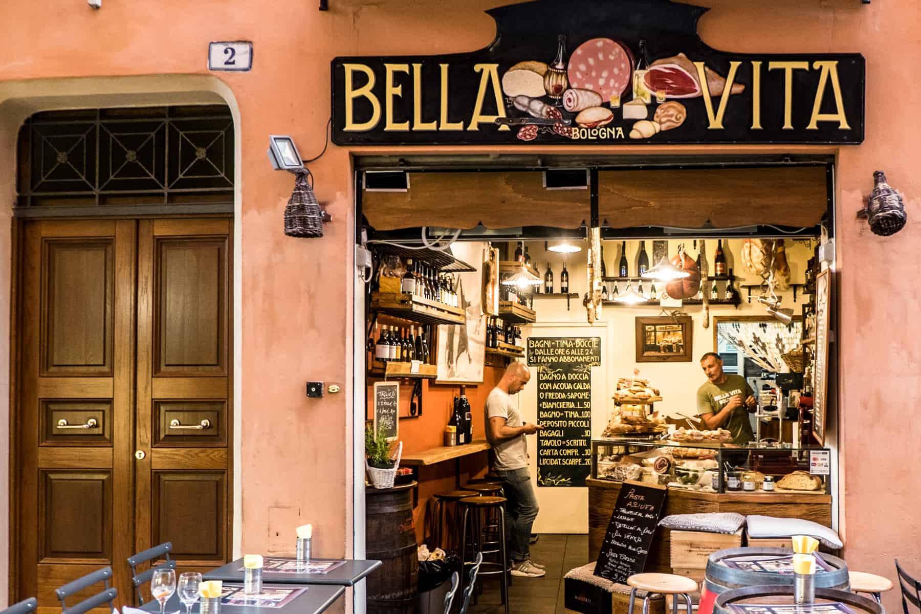 Two men in a small bar in Bologna. The sign says "Bella Vita" with pictures of meet, cheeses and breads. Tables and chairs stand outside the bar. 