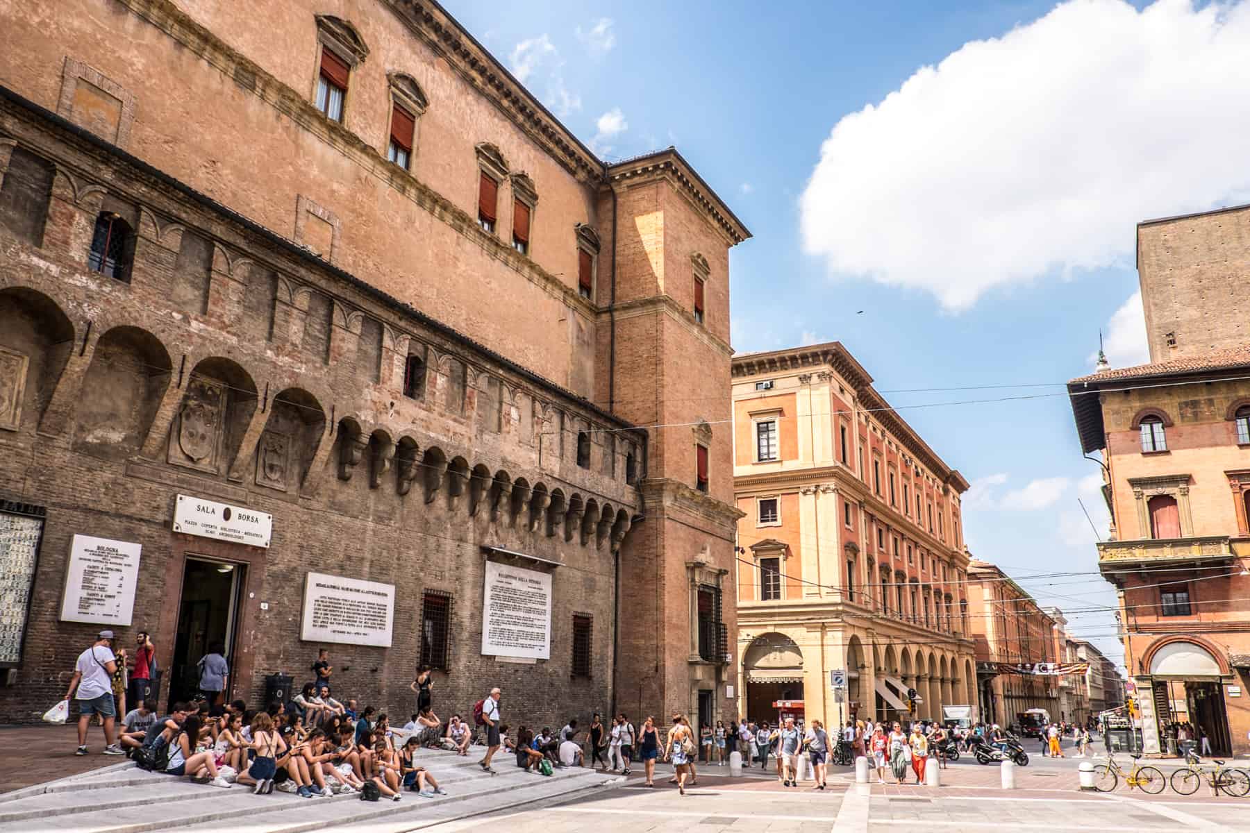 People sitting on the stairs below the white signs on a tall, brown and orange stone medieval building in Bologna. 