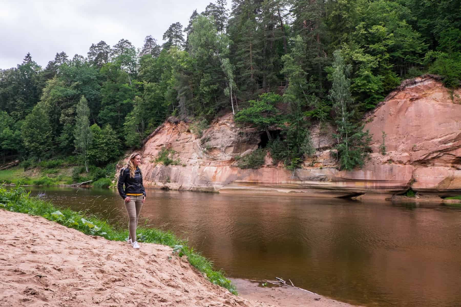 A woman in khaki pants and a black jacket stands on a sandy riverbank looking towards an ochre red cave under a green forest.