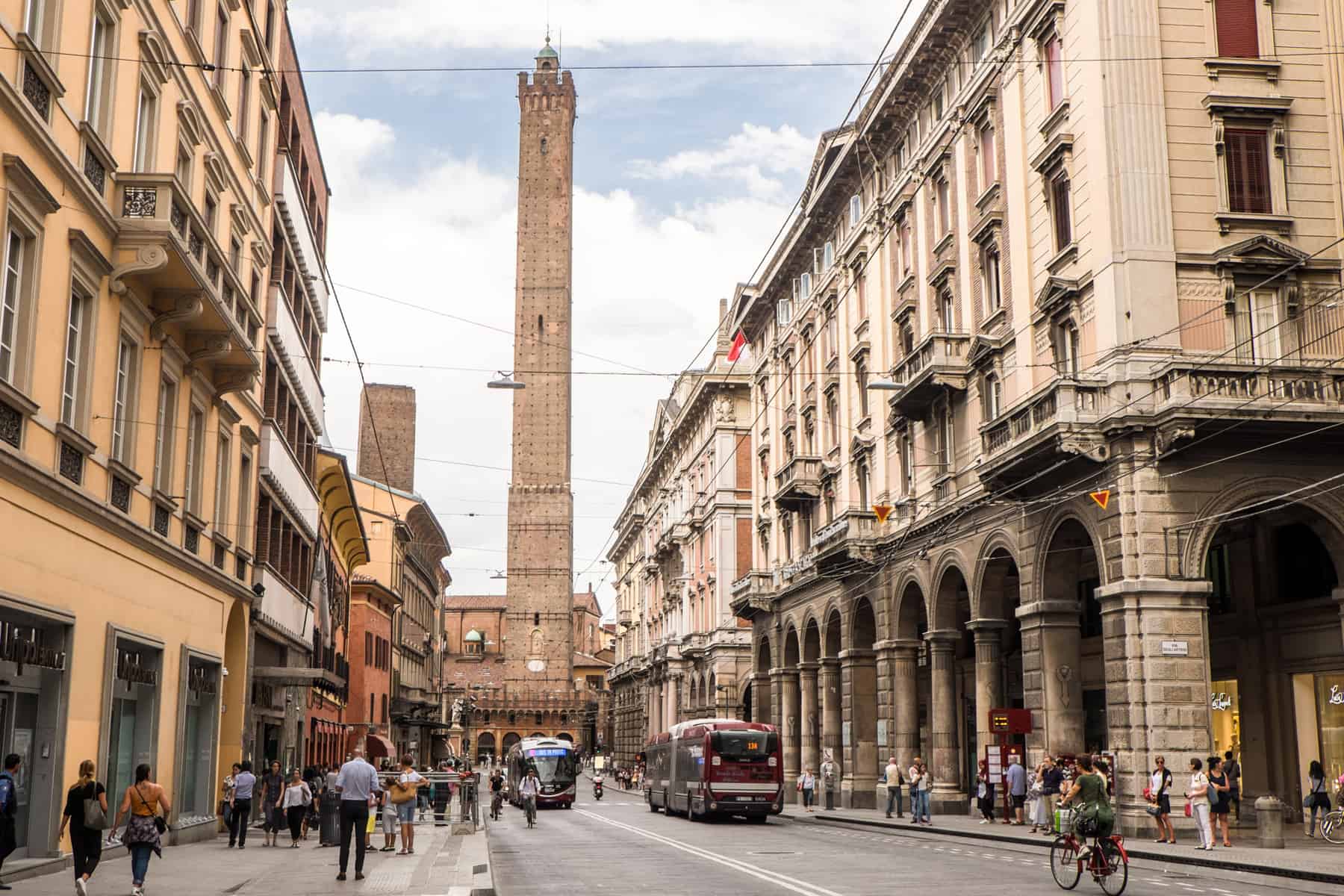 A tall medieval stone tower stands at the end of a wide street in between row of beige buildings, and dominates the skyline of Bologna.