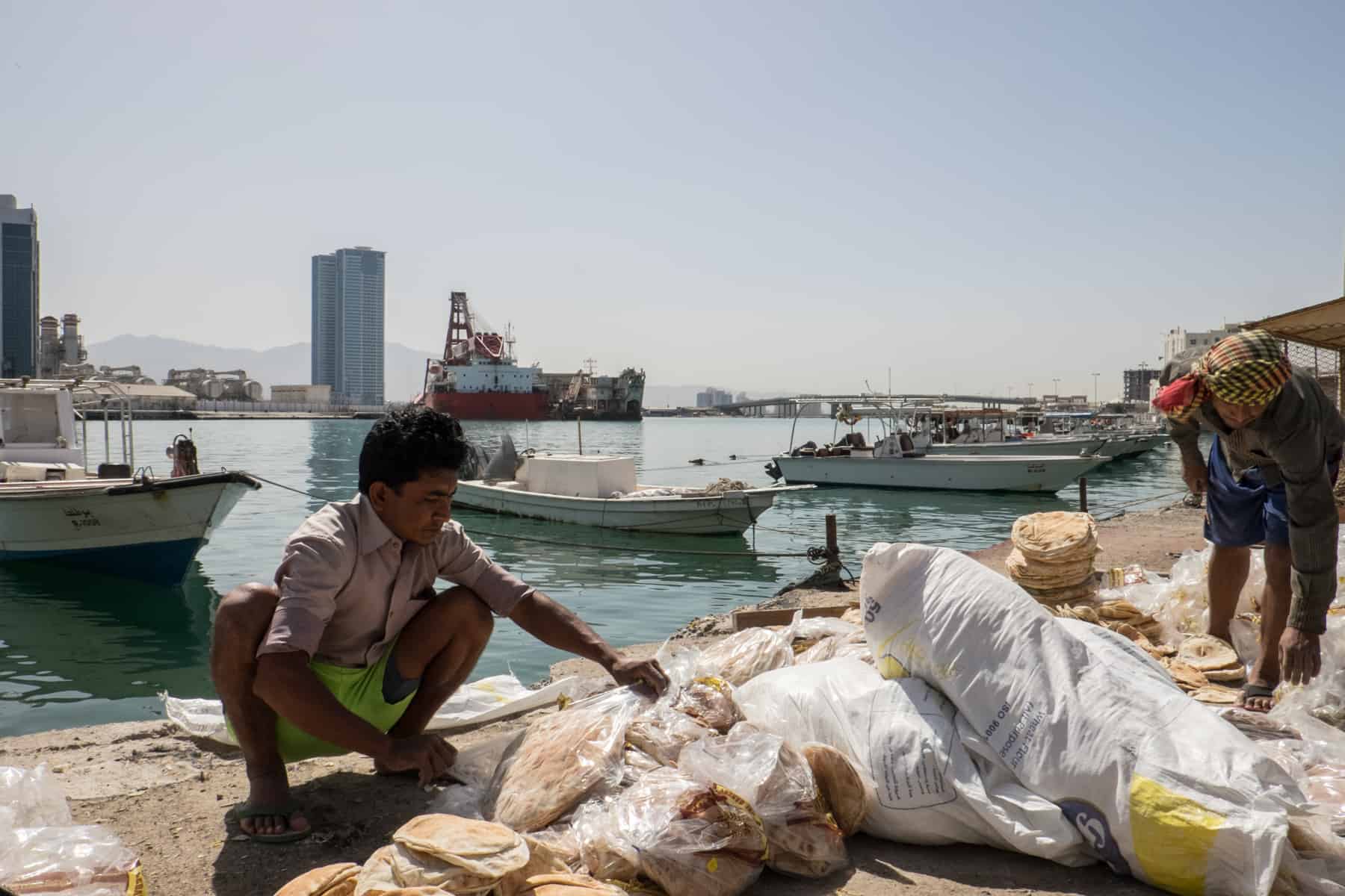 A man, squatting, sells flat bread from bags next to the waterfront and port area in Ras Al Khaimah, UAE