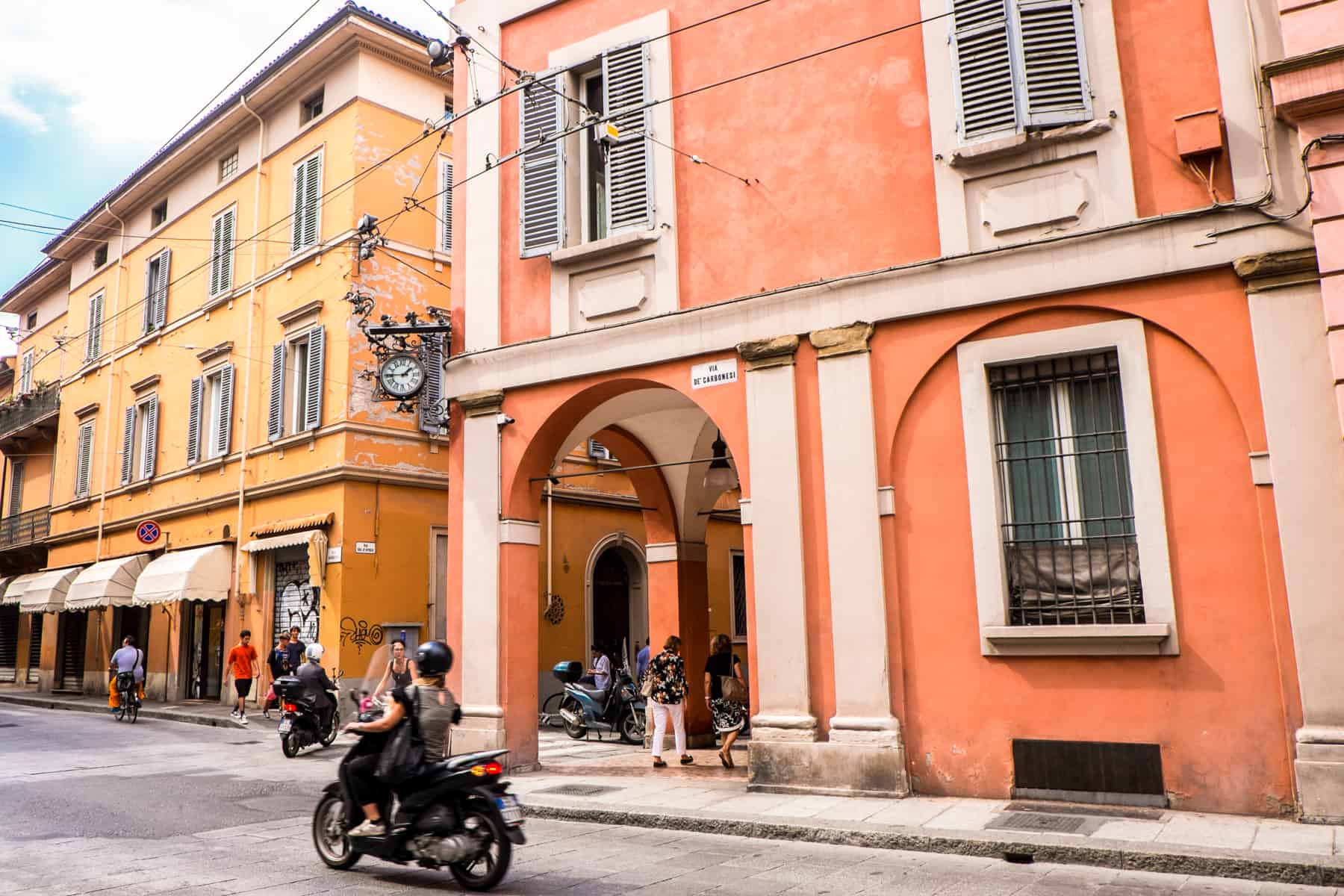 A woman on a motorbike ride past an orange building in Bologna, which stands next to a similar structure painted in yellow. 