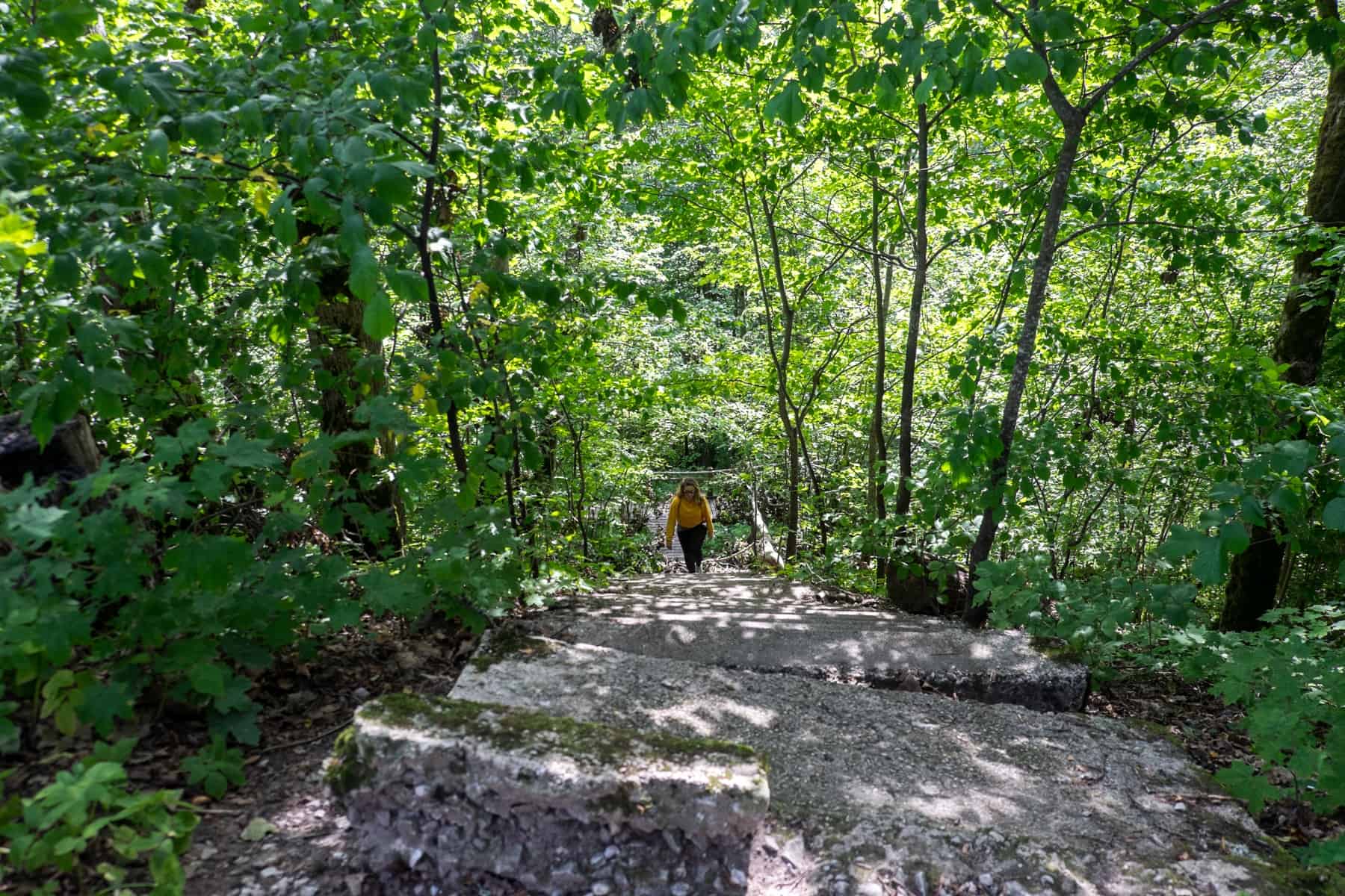 A woman in a yellow jumper walks up stone steps in a green forest in Sigulda, Latvia.