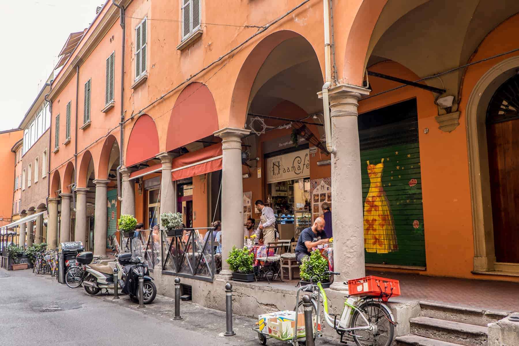 People sit outside a cafe, set within the archway corridor of a long, orange building in Bologna, Italia.