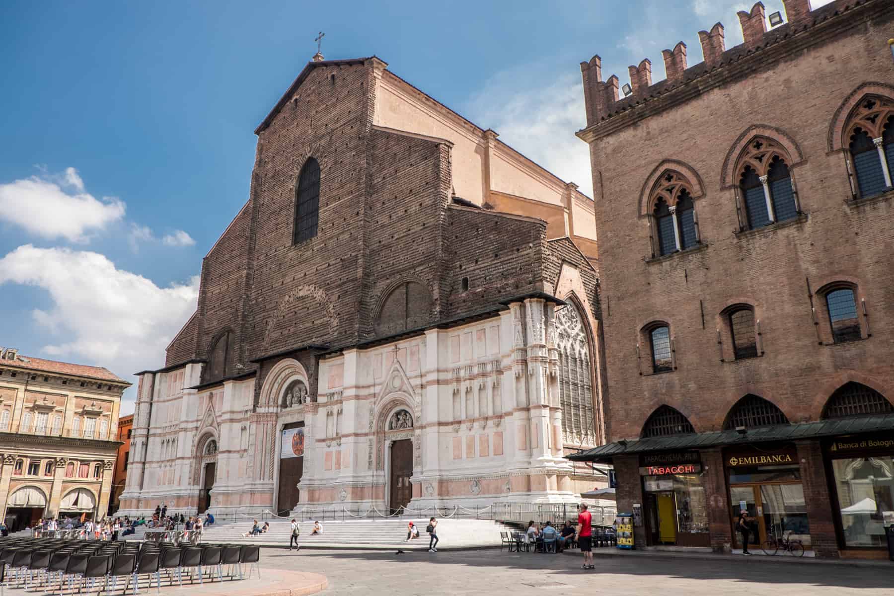A church in Bologna city with a white and salmon pink stone bottom half and a brown stone top half. To the right is a brown, medieval building with a castle-like turret roof. 