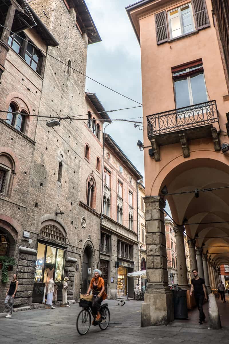 A woman rides a bike down a street in Bologna with brown medieval stone buildings on one side and the archway portico corridor buildings on the other. 