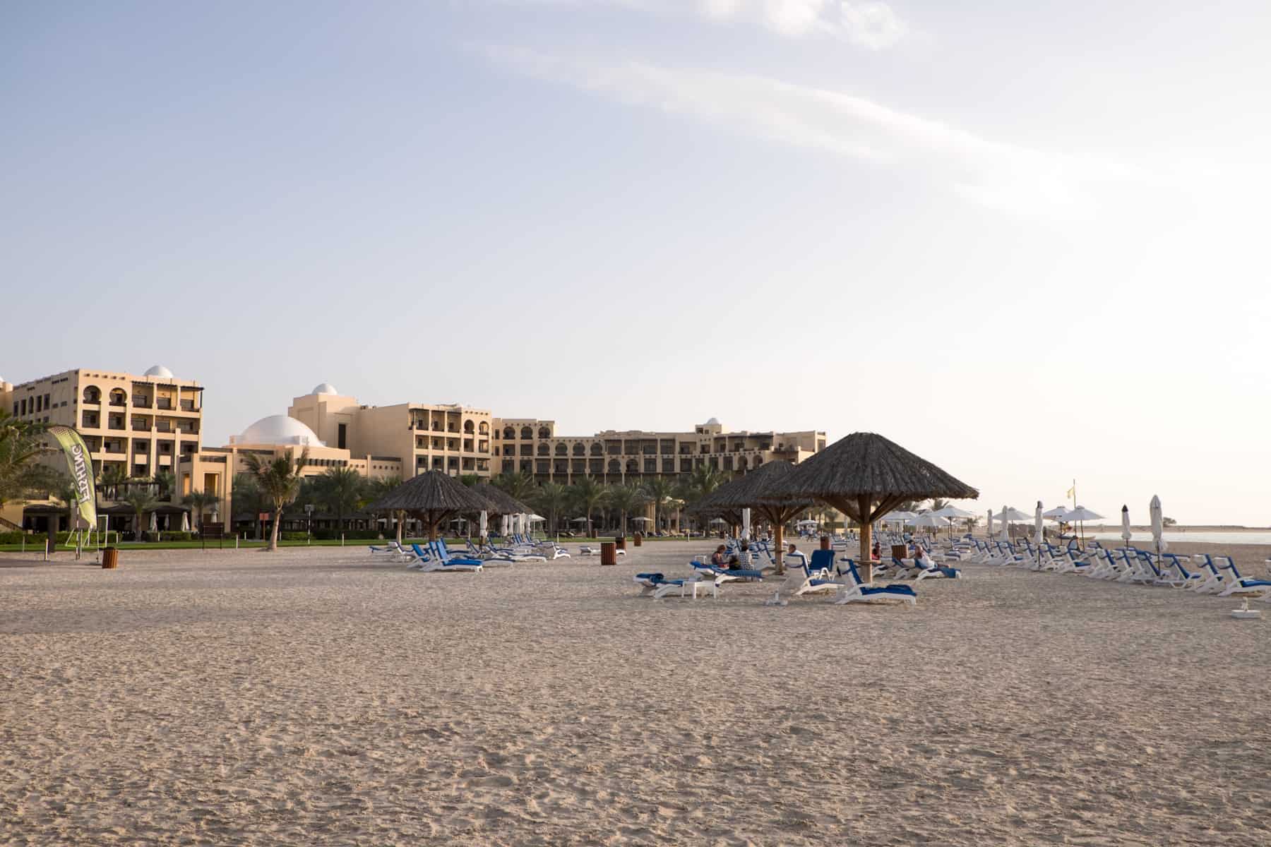 A sand coloured, low-rise hotel complex next to a sandy beach with wicker umbrella shades and blue covered sun loungers