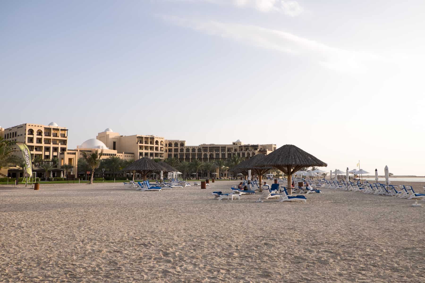 A sand coloured, low-rise hotel complex next to a sandy beach with wicker umbrella shades and blue covered sun loungers