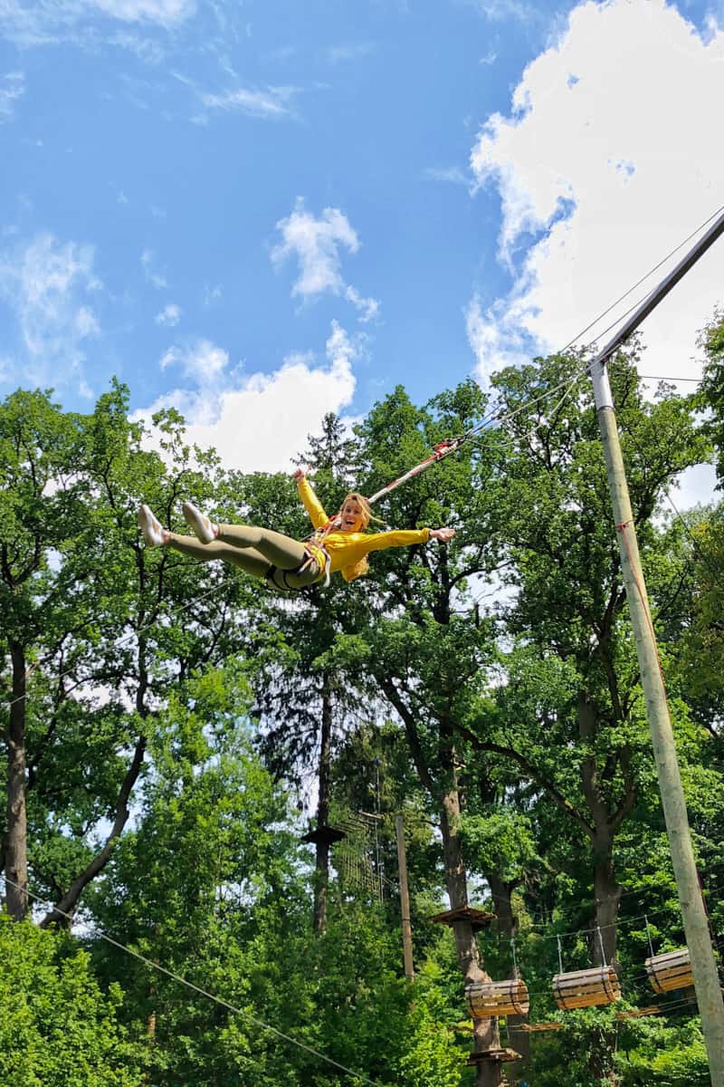 A woman in green jeans and yellow jacket is suspended mid air against a backdrop of trees, happily screaming on a cataput line at an adventure park.