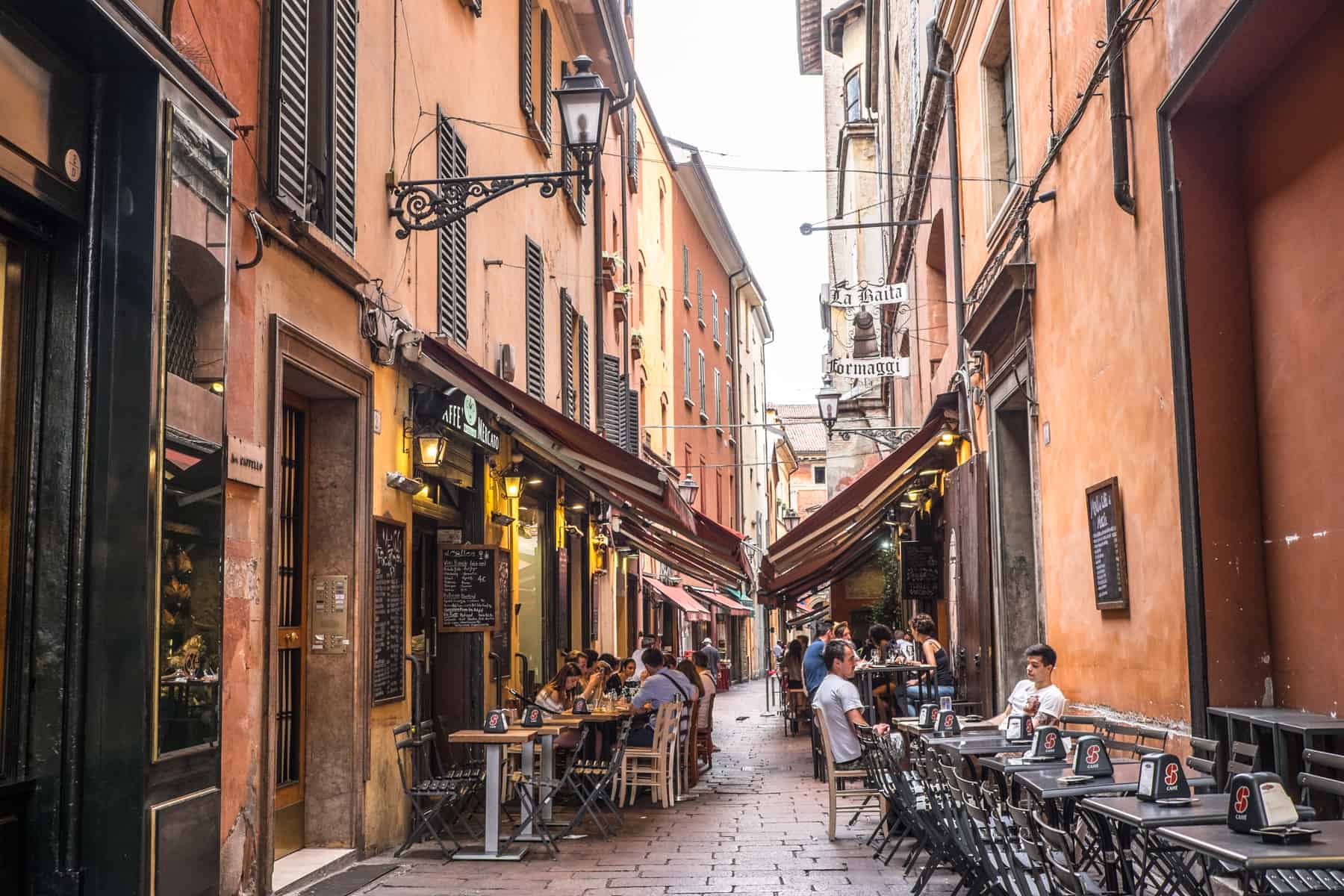 A narrow street in Bologna Italy lined with orange building and filled with tables and chairs and people dining al fresco. 