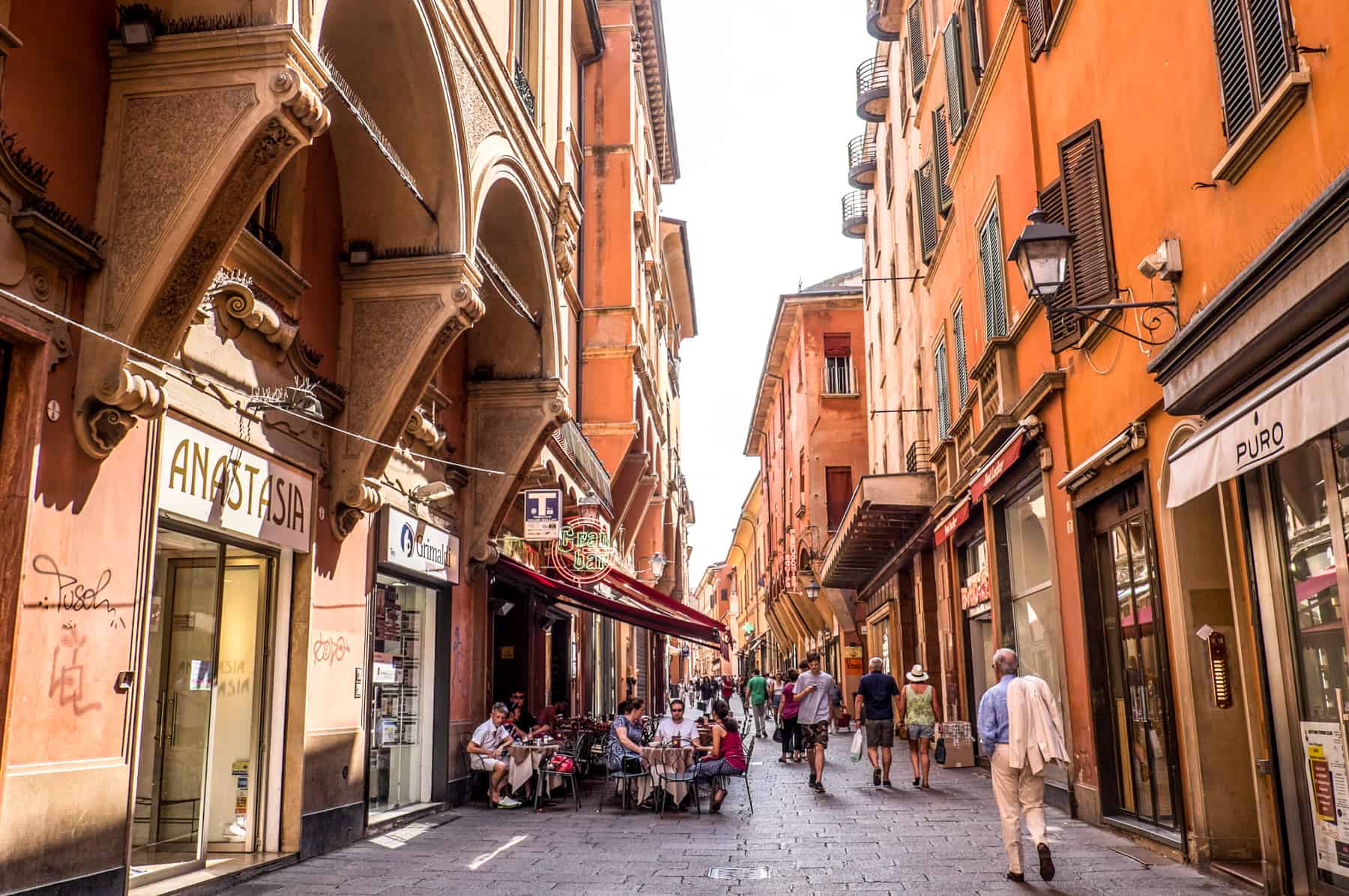 A busy pedestrian street in Bologna Italy lined with orange buildings and shop fronts. People are walking past a small group sitting around tables at an outdoorscafe. 